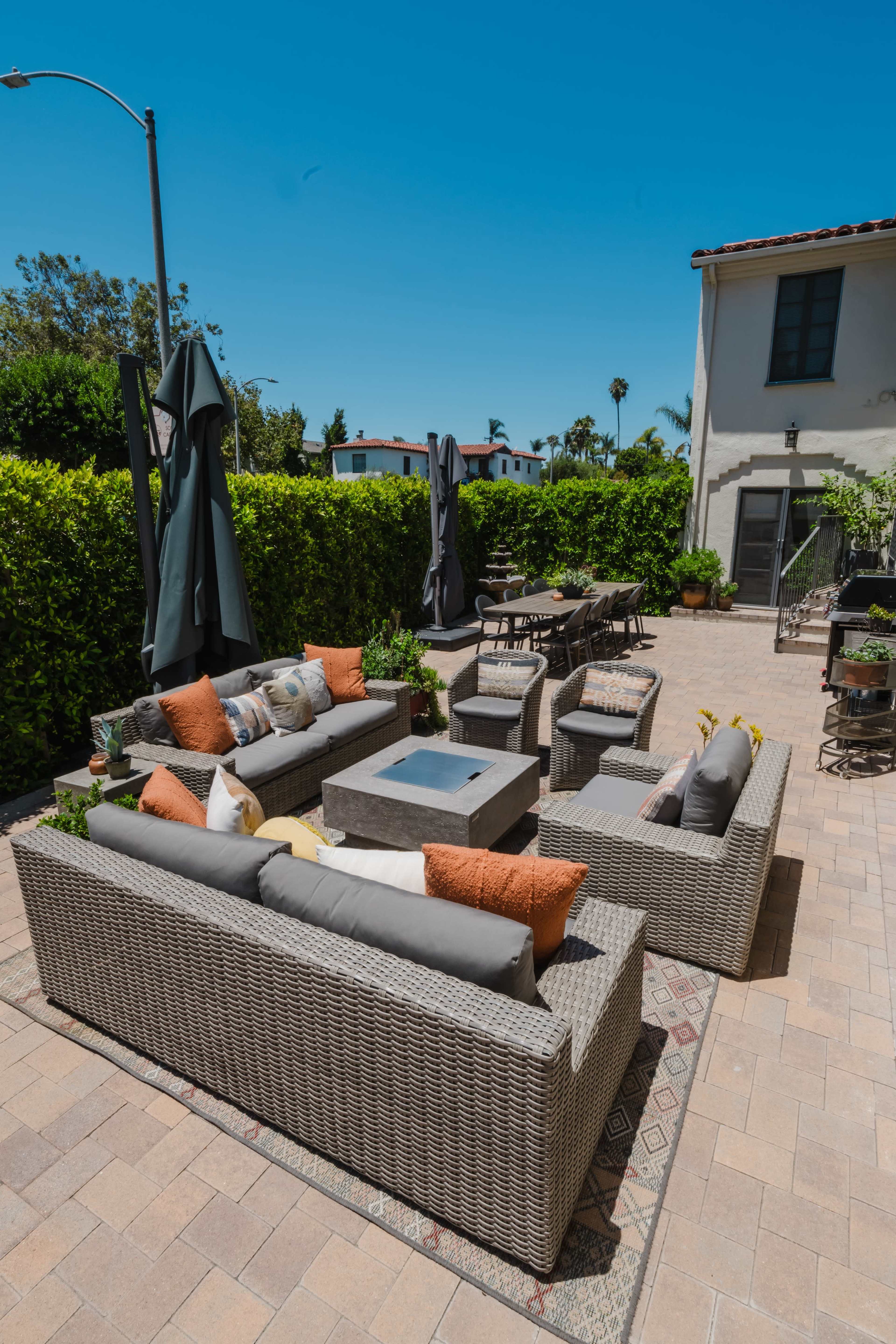 The image shows a patio area with a set of outdoor furniture, including a sectional sofa and chairs arranged around a central fire pit, surrounded by greenery.