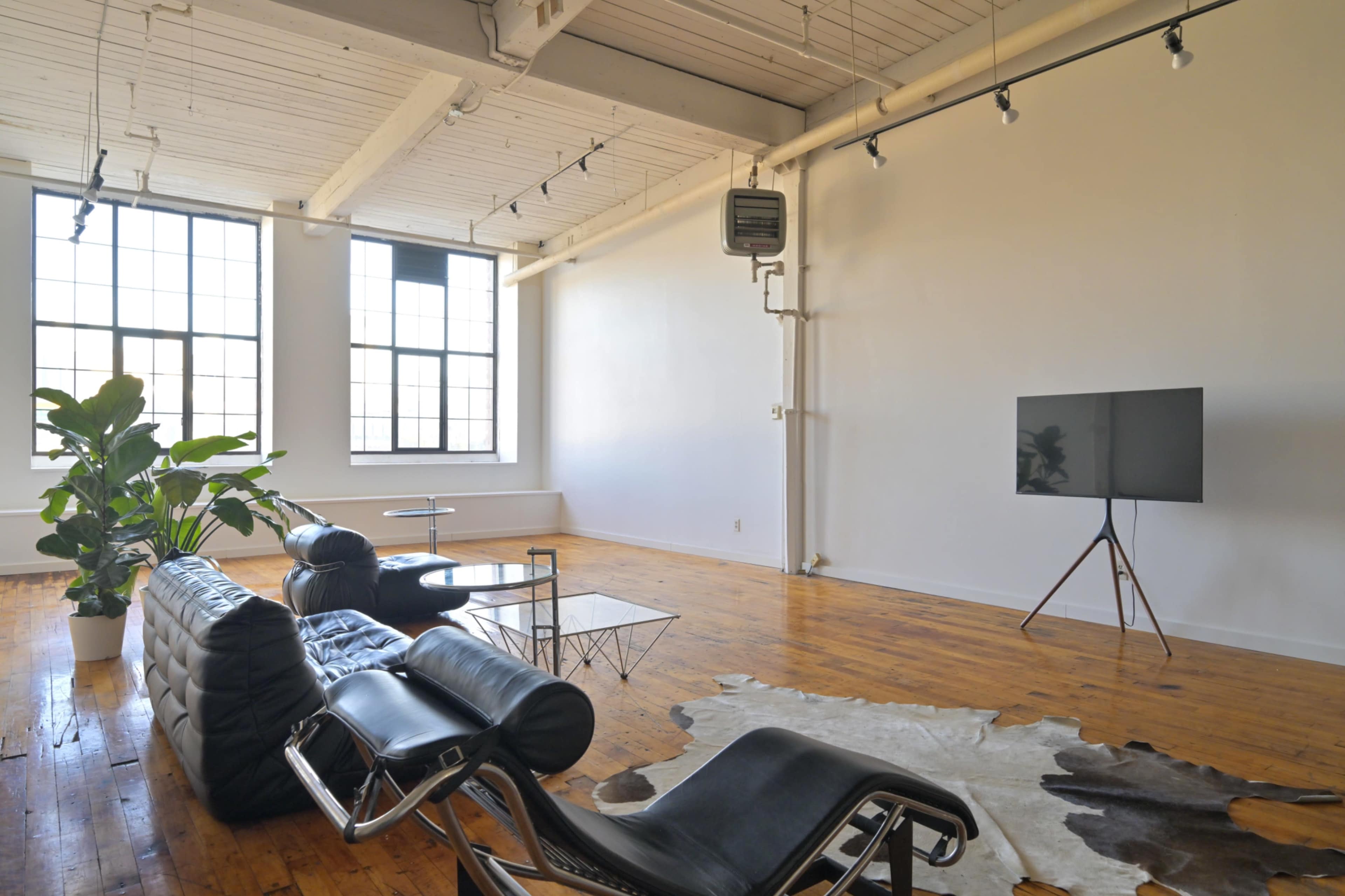 The image shows a spacious, minimalist living room featuring large windows, a leather couch, a glass coffee table, a television on a stand, and a potted plant.