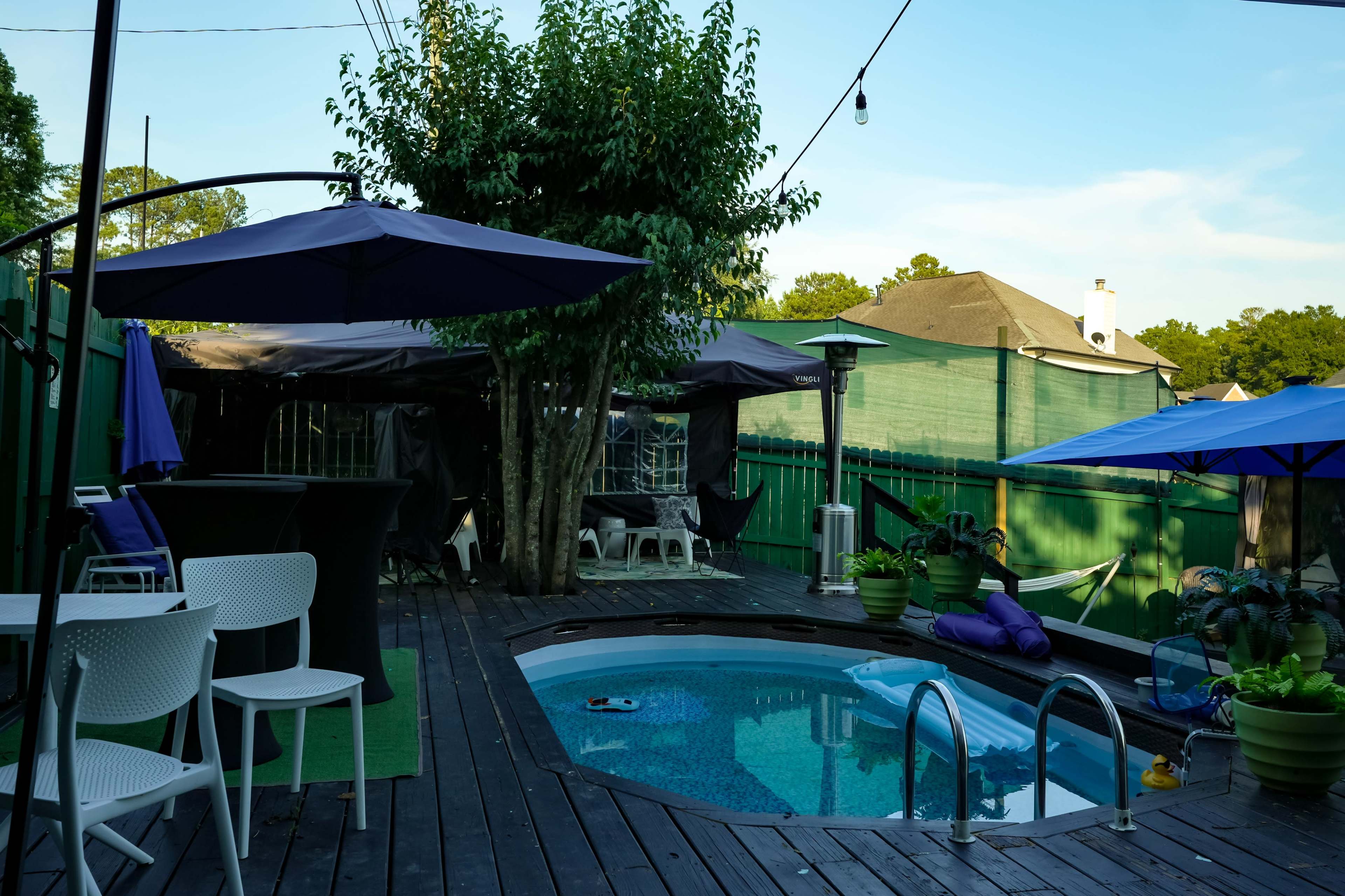A wooden deck features a small pool surrounded by patio furniture and several umbrellas, with greenery and a shaded area in the background.