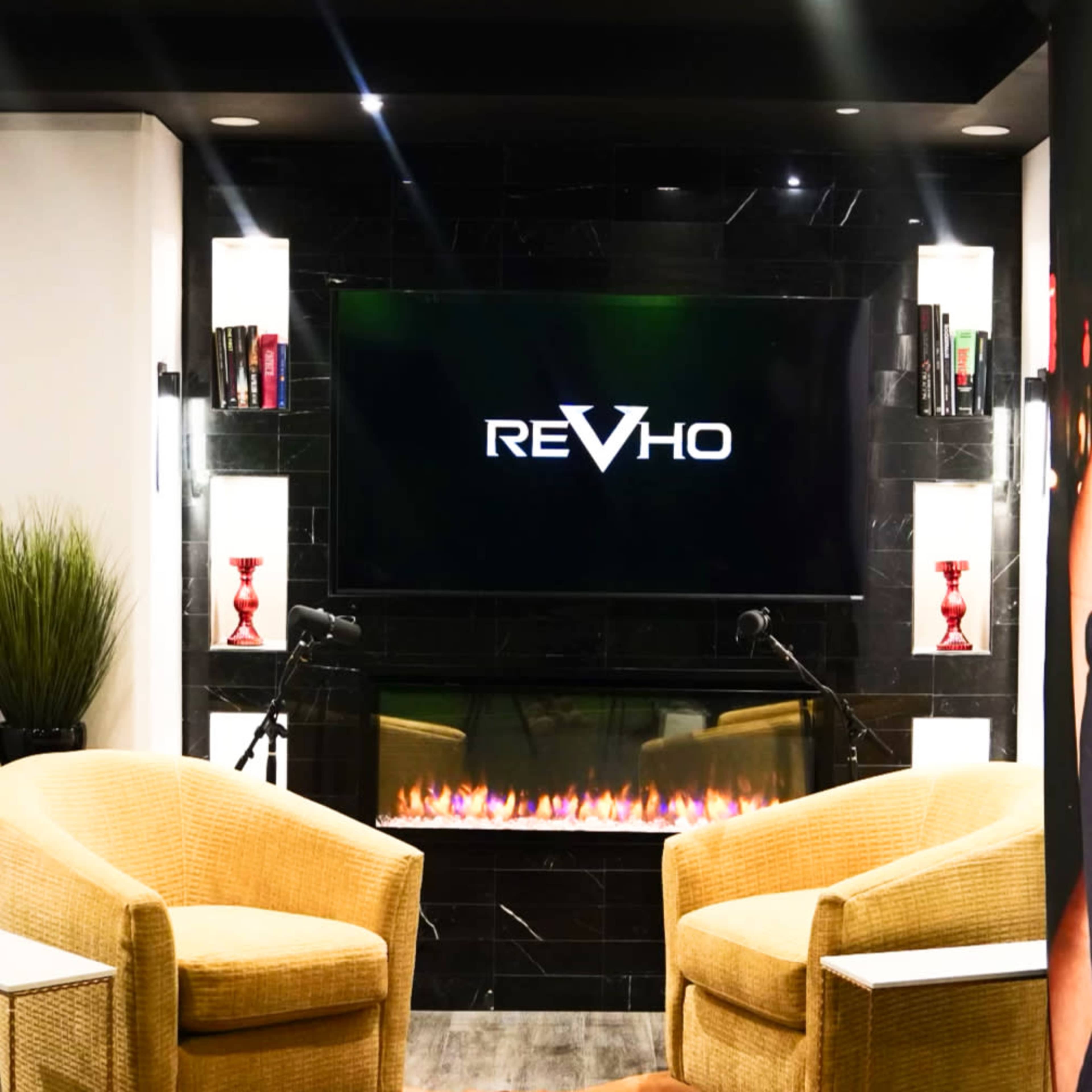 The image shows a modern lounge area with two yellow armchairs, a wall-mounted television displaying the logo "REVHO," and an electric fireplace beneath it.