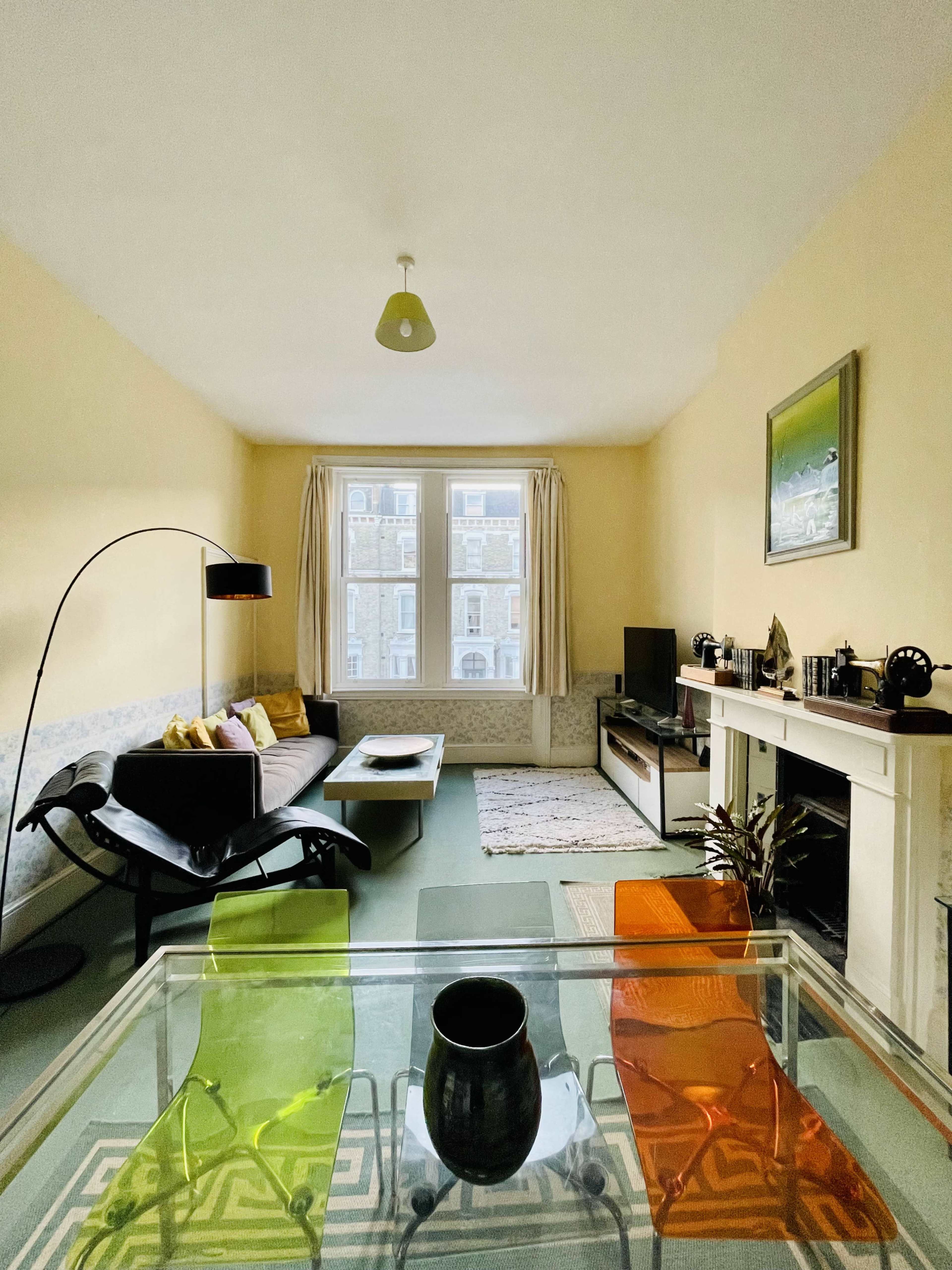 The living room features a sofa, a glass coffee table with colorful chairs, a television, and a fireplace, with large windows allowing natural light to enter.