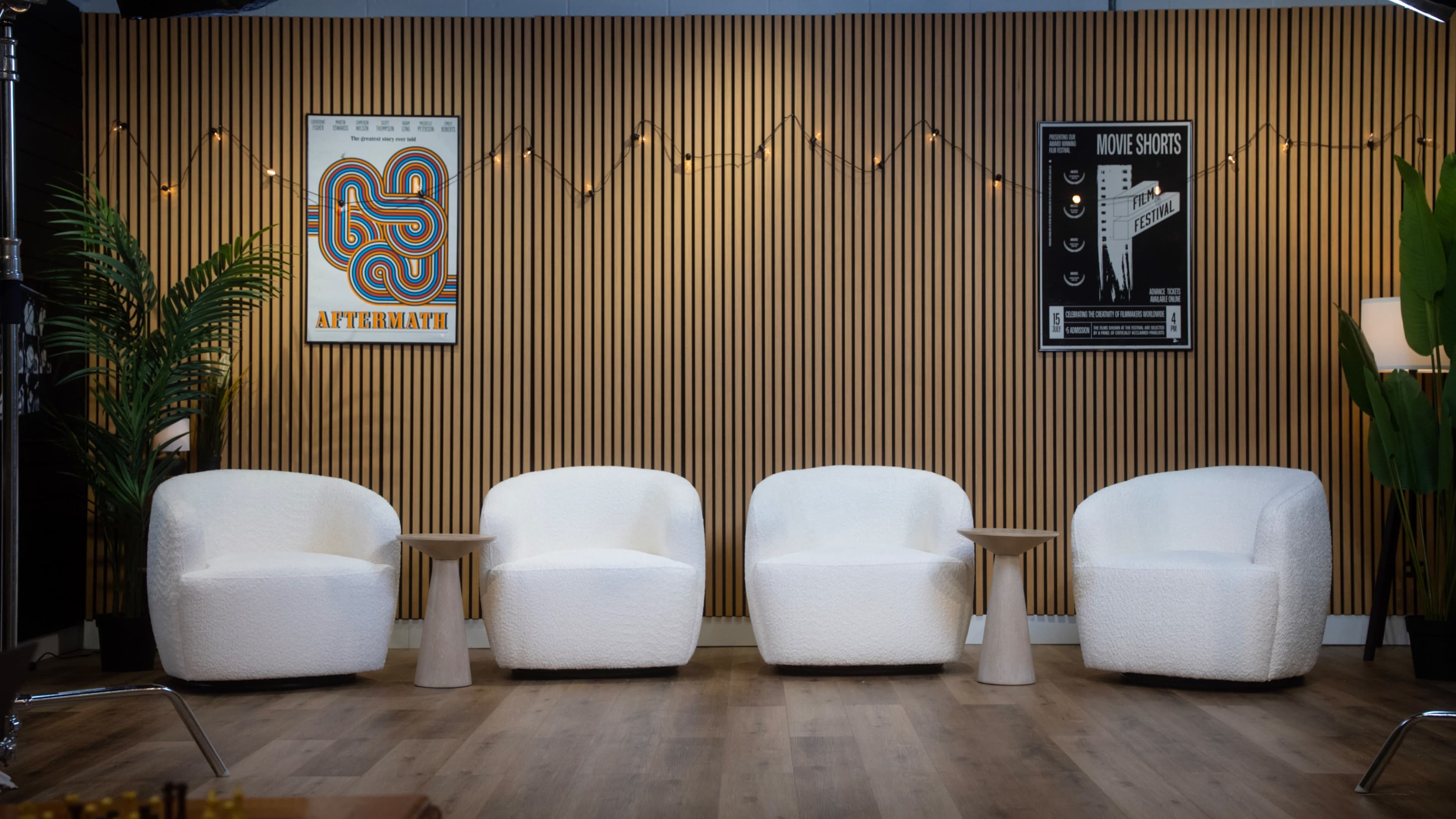 The image shows a modern interior with four white, rounded chairs set around small wooden tables against a backdrop of vertical wood paneling and decorative posters.