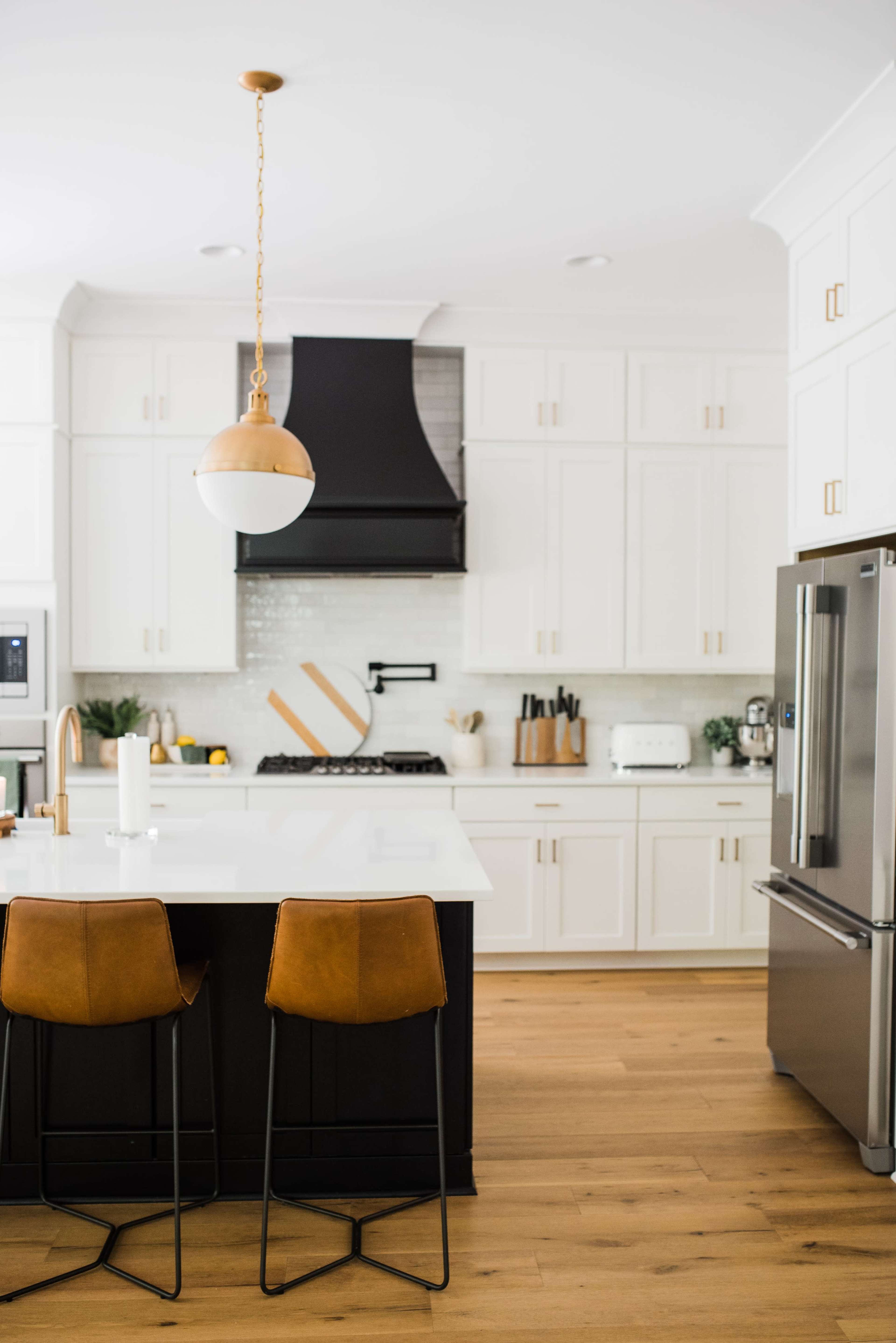 A modern kitchen features white cabinetry, a black hood above the stove, a large island with two brown bar stools, and stainless steel appliances.