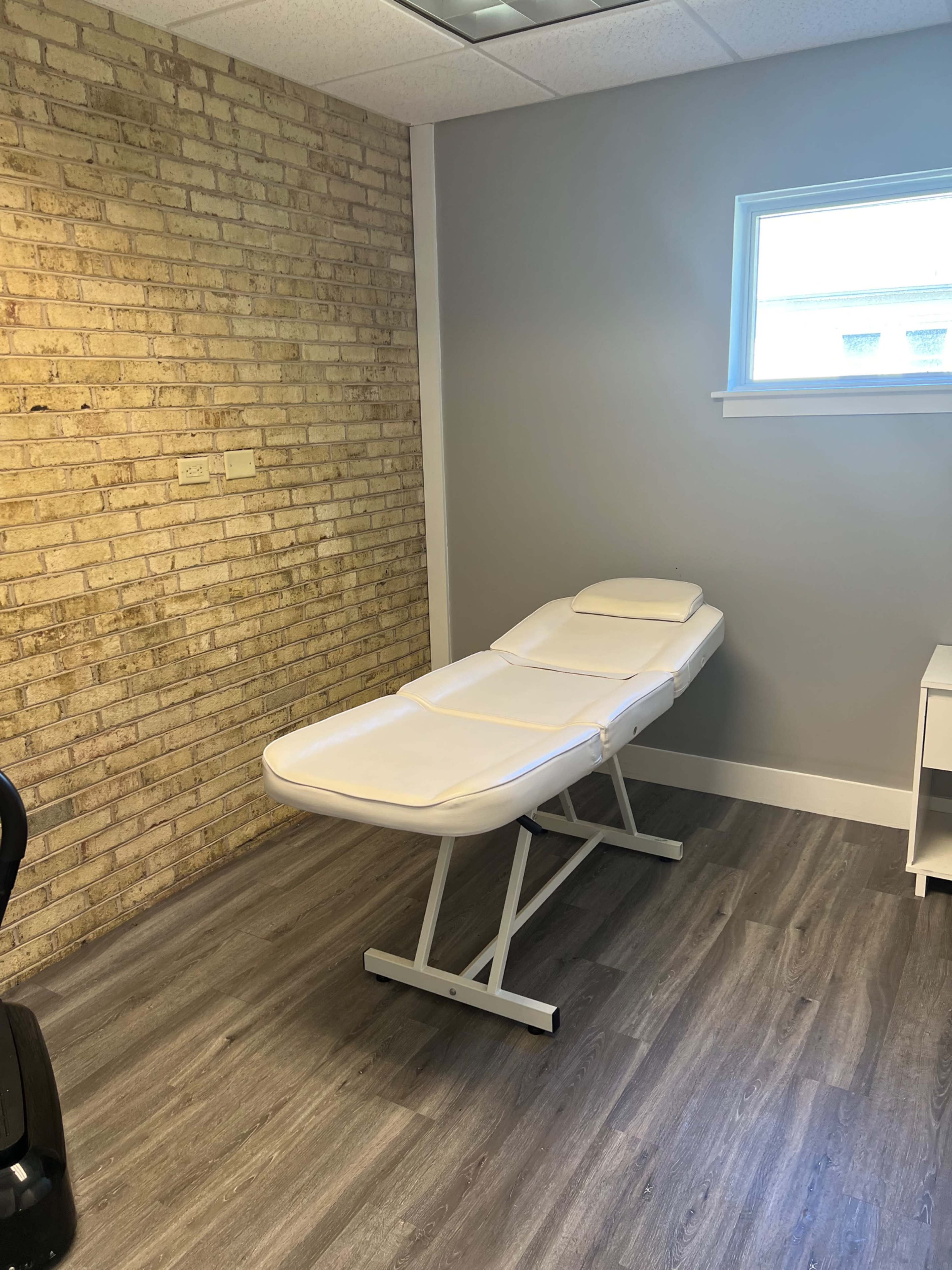 A clean treatment room featuring a white examination table against a brick wall and a window.