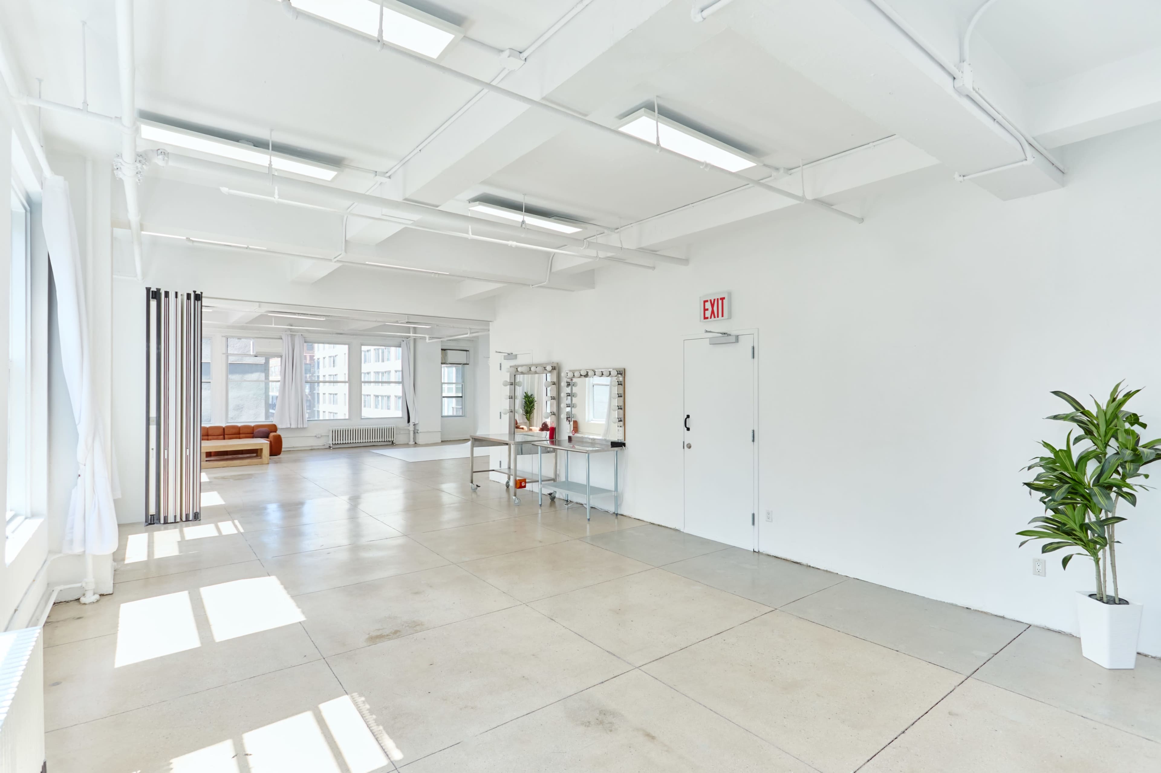 The image shows a spacious, well-lit interior with minimal furniture, featuring large windows, a small table with a mirror, and a plant in one corner.