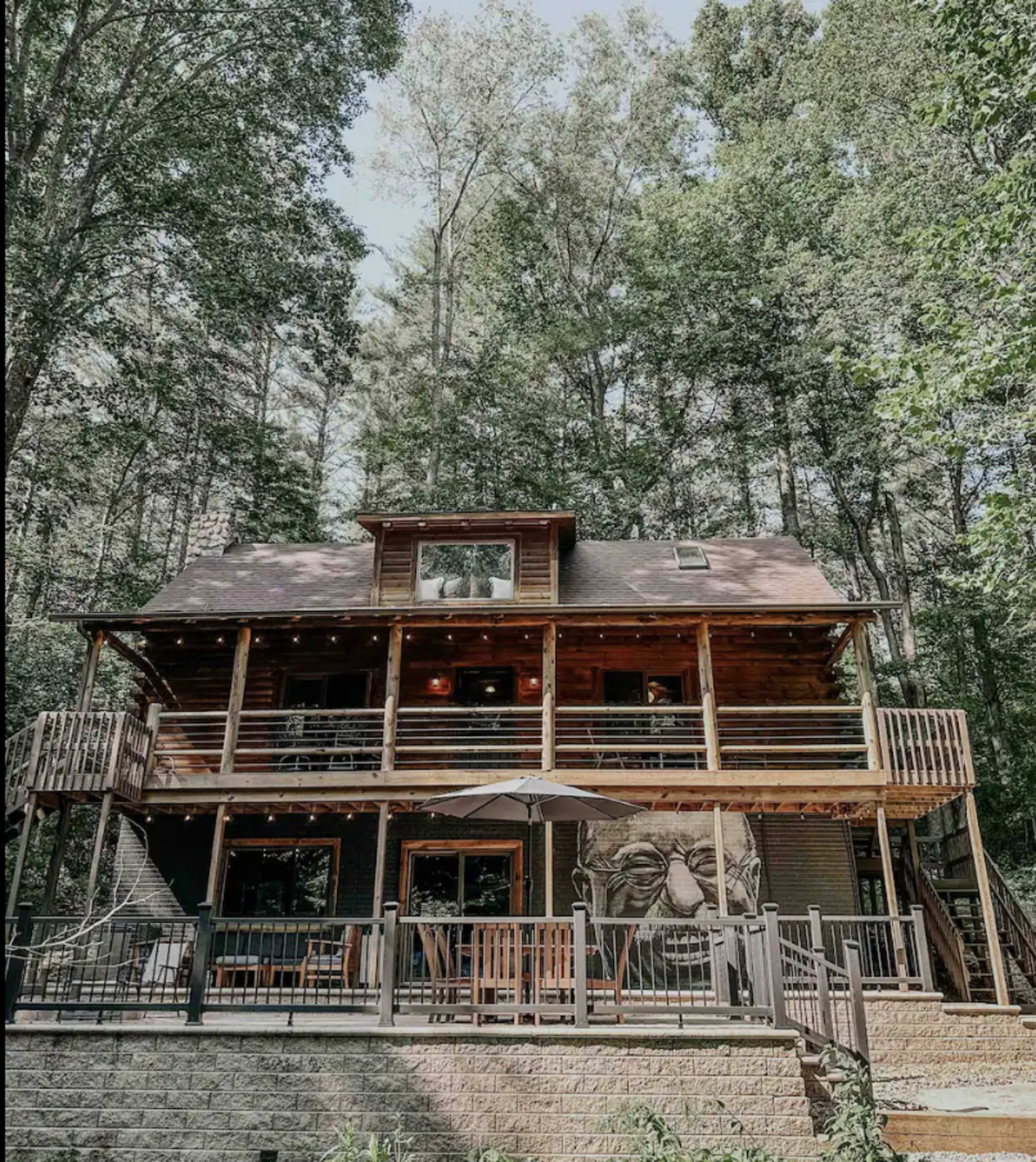 A large wooden cabin with multiple decks and a carved face feature stands surrounded by tall trees.