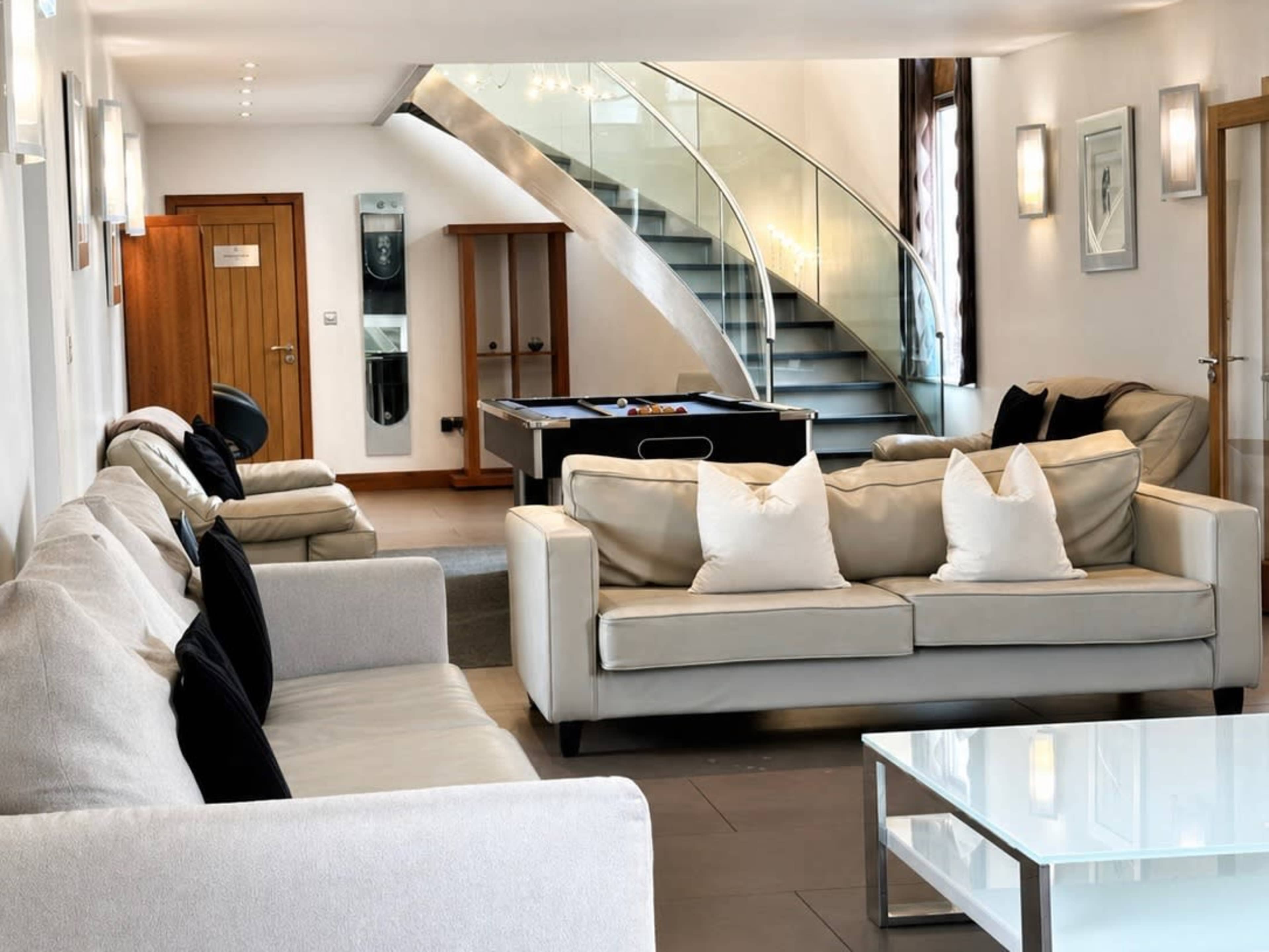 A modern lounge area features light-colored sofas with black and white pillows, a glass coffee table, and a staircase in the background.