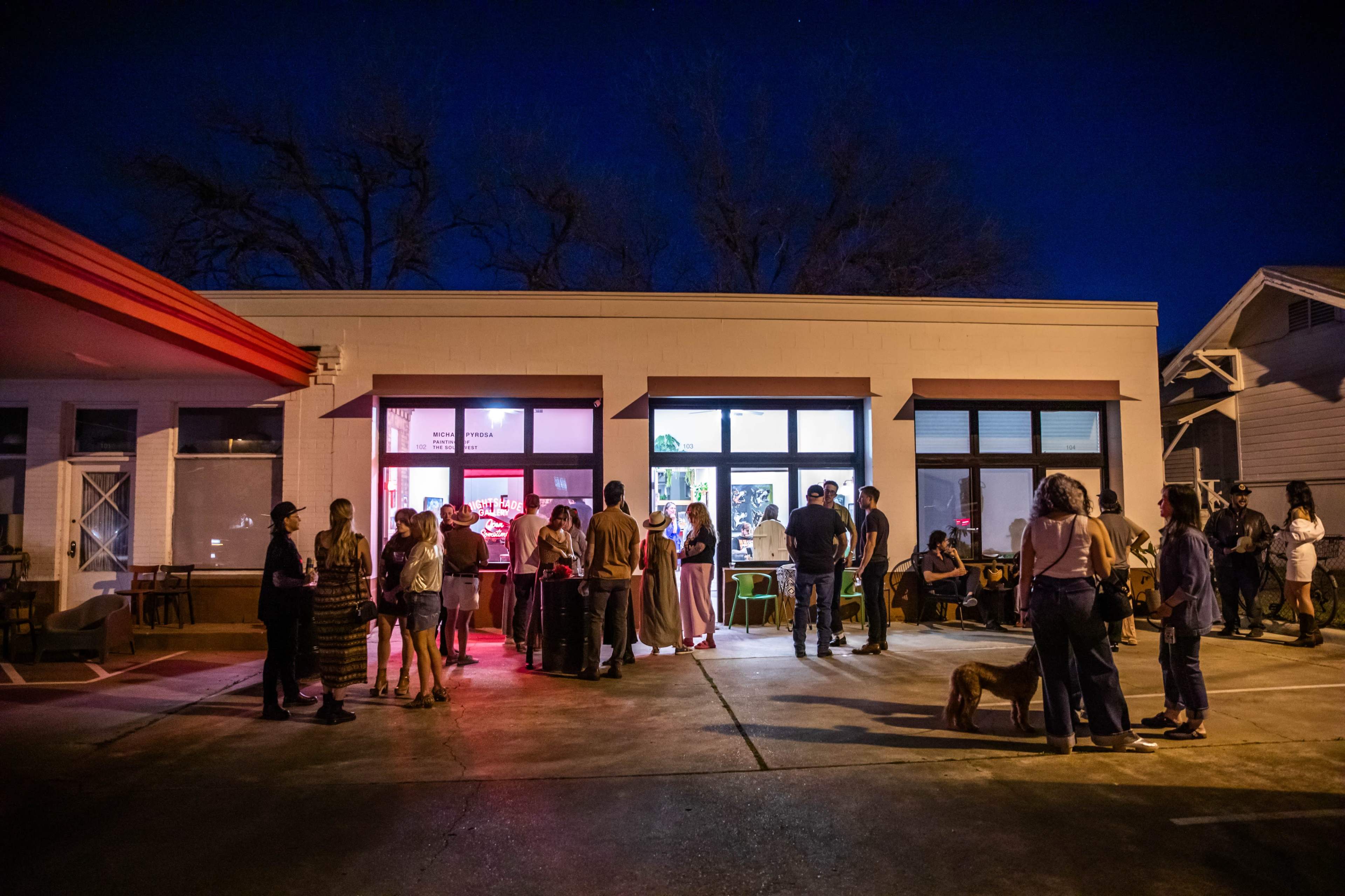 A crowd of people gathers outside a brightly lit building at night, with some standing and others seated in a casual outdoor setting.