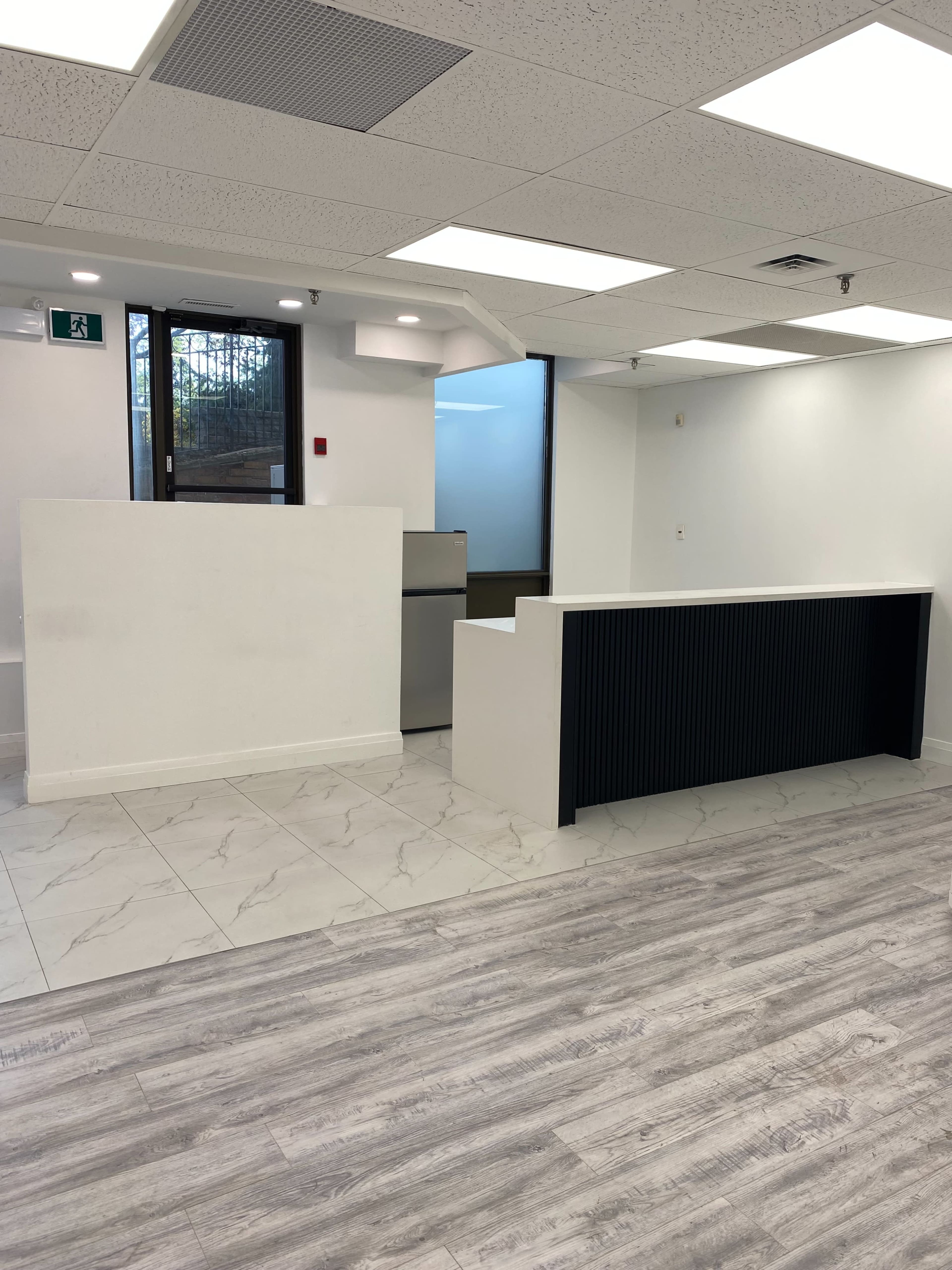 The image shows a spacious office interior featuring a reception area with two counters, one white and one black, and large windows allowing natural light.