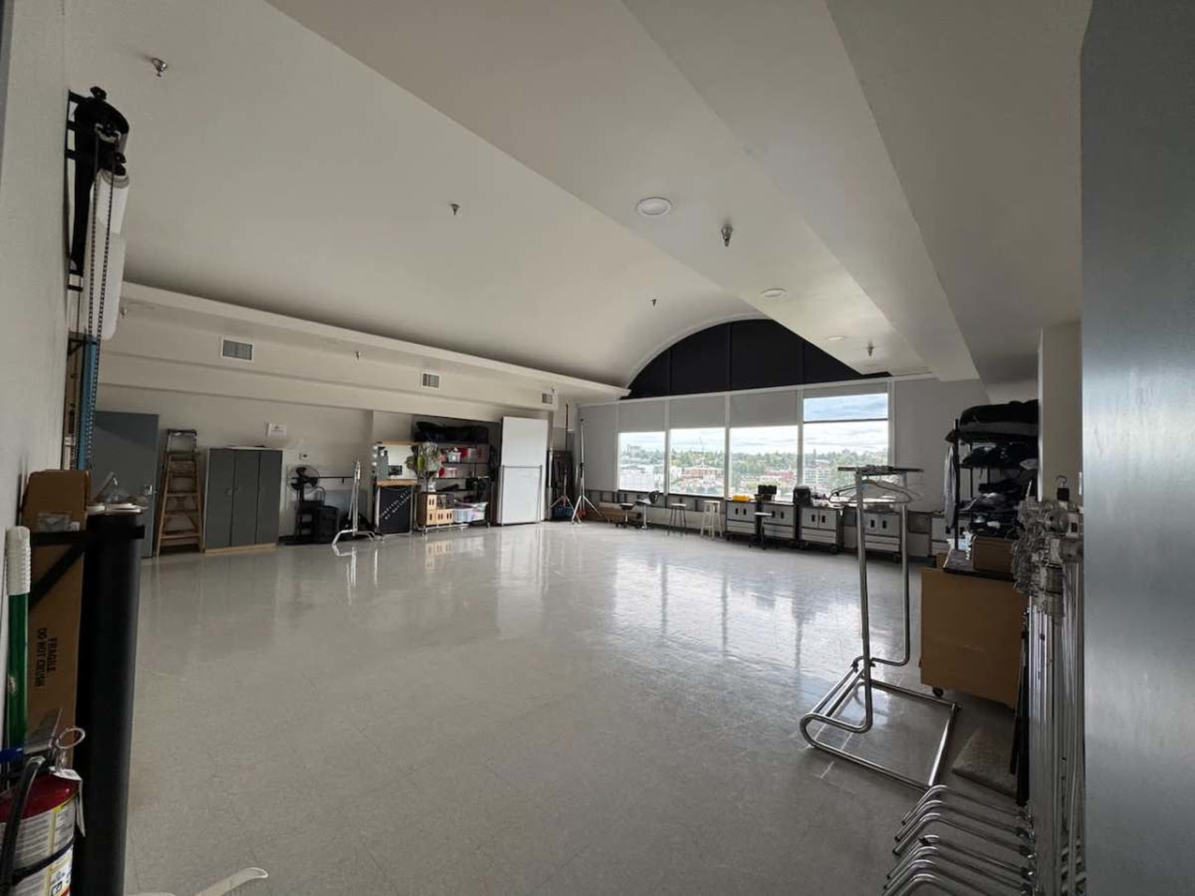 The image shows a spacious, empty room with large windows, boxes, and storage units along the walls.