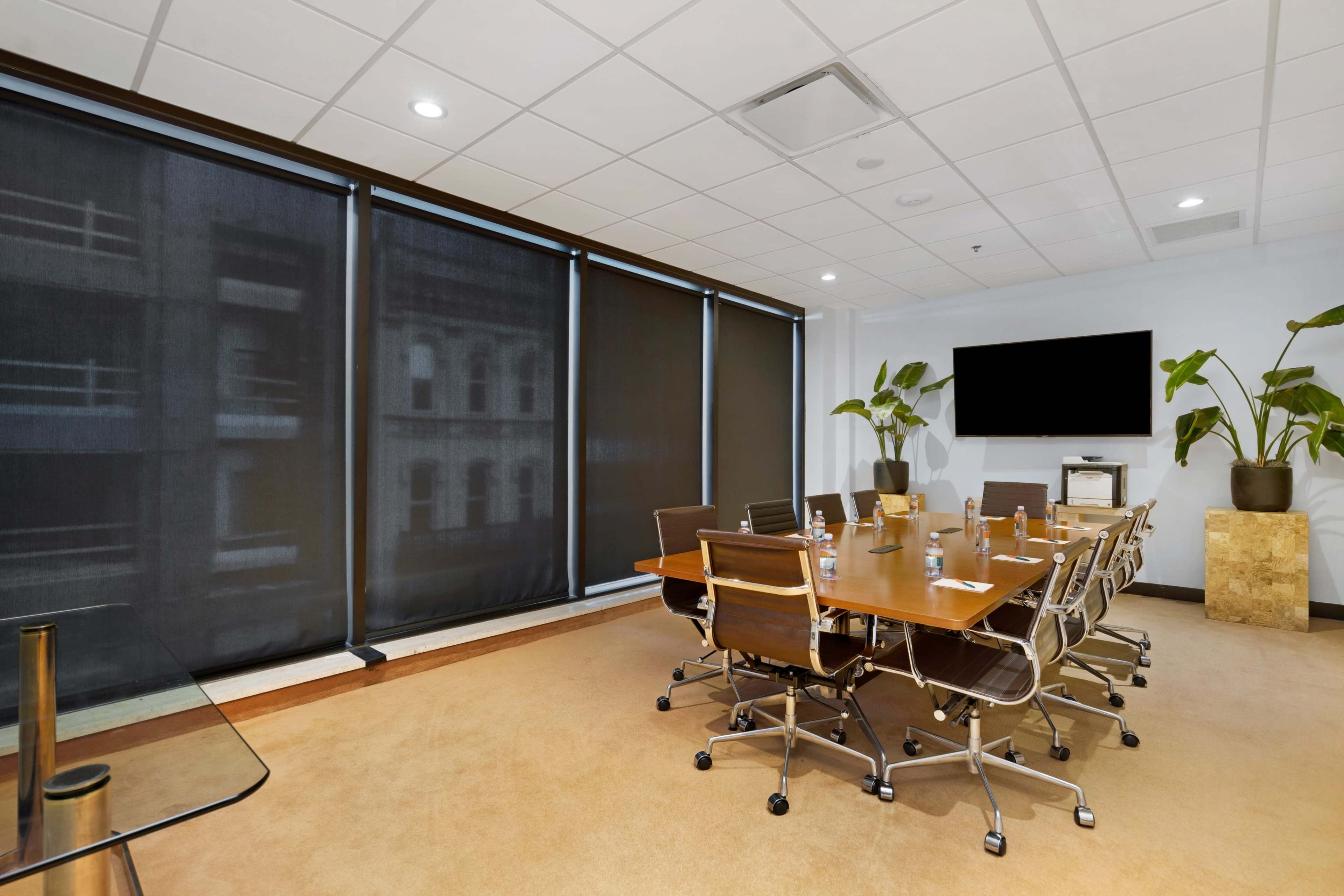 A modern conference room features a large table surrounded by ergonomic chairs, with dark window shades and a wall-mounted screen.
