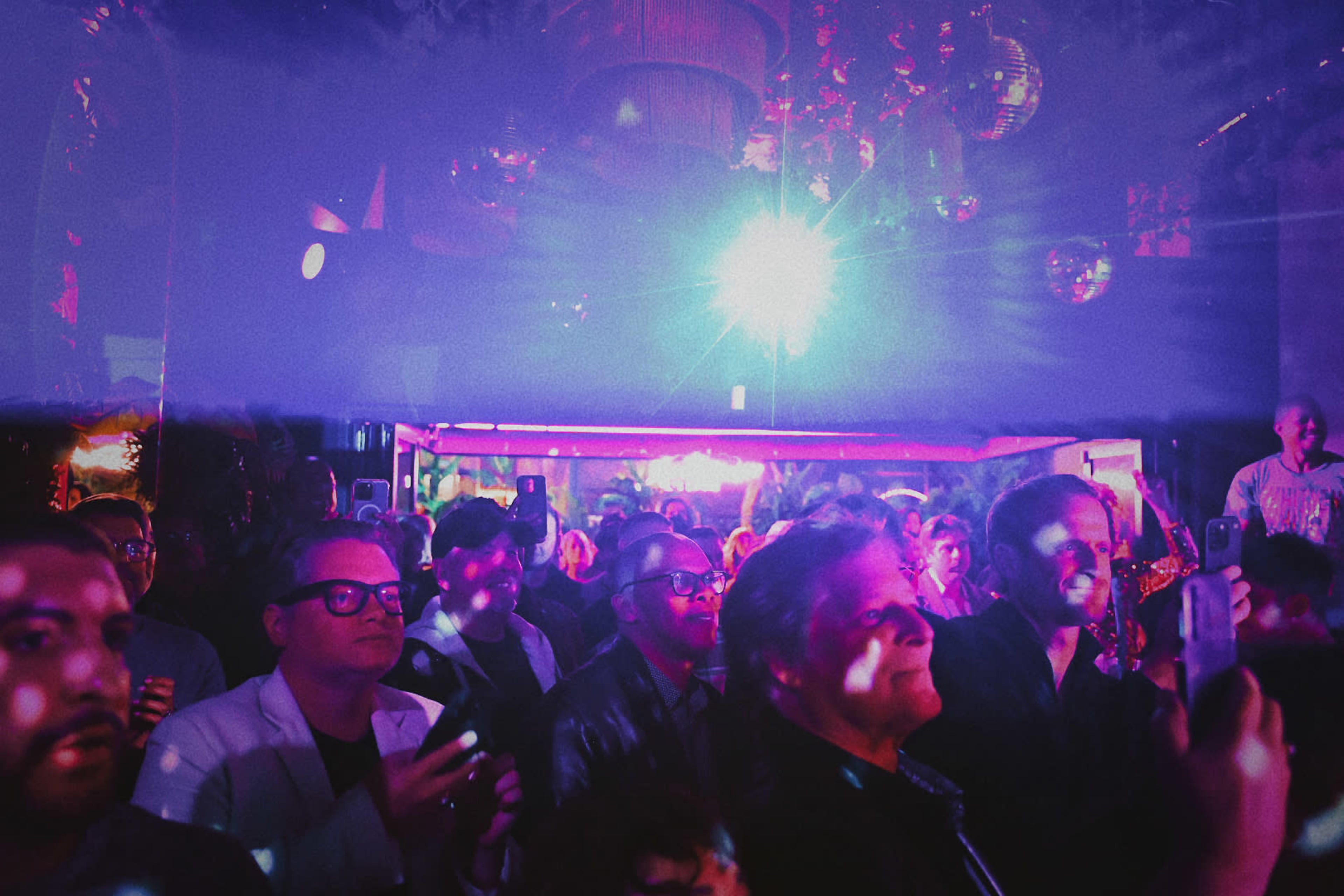 A crowd of people stands in a dimly lit venue, capturing the performance with their phones while a colorful light beam shines overhead.