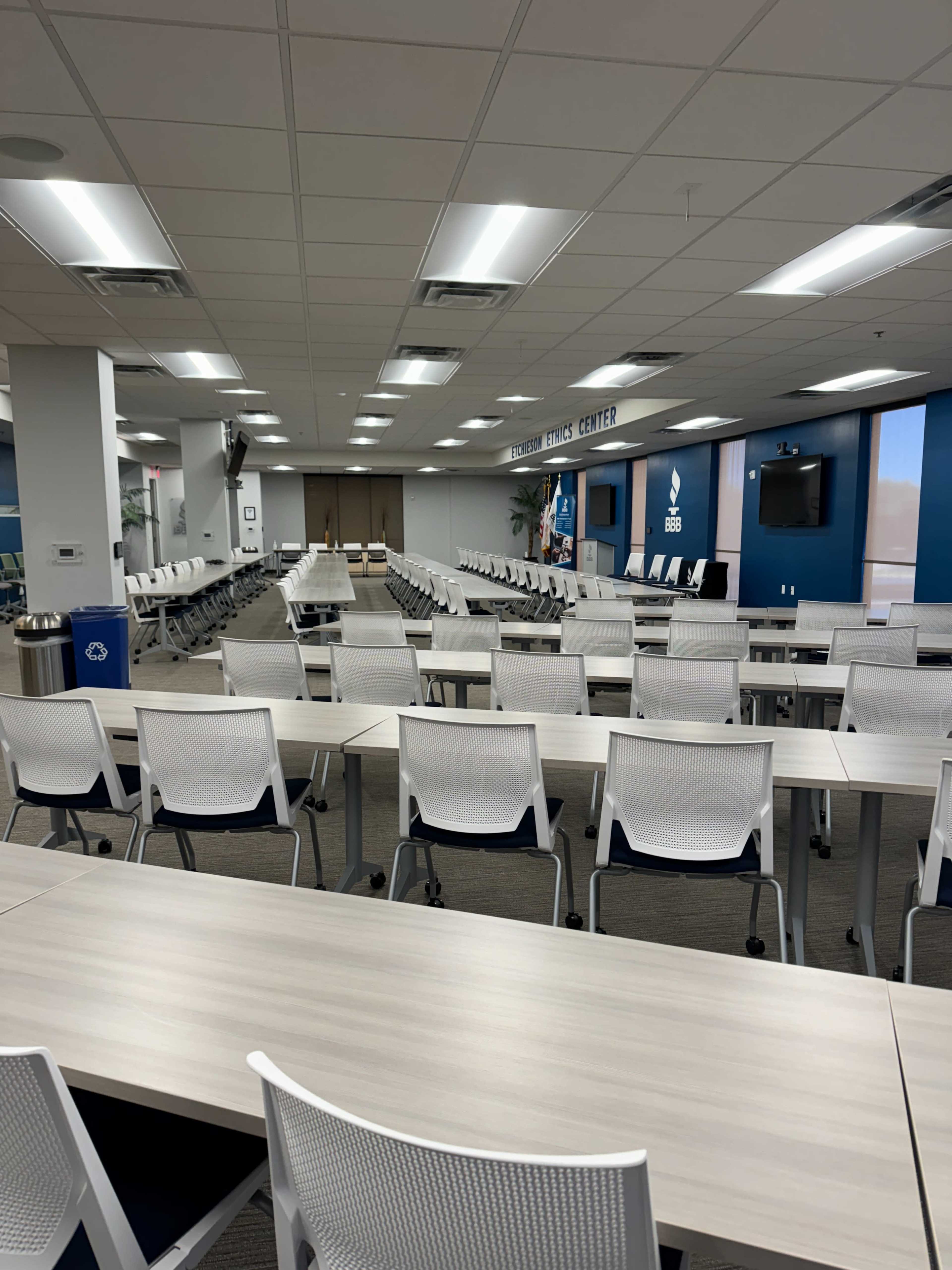 The image shows a conference room with rows of tables and chairs set up for a meeting, featuring a large screen and a podium in the front.