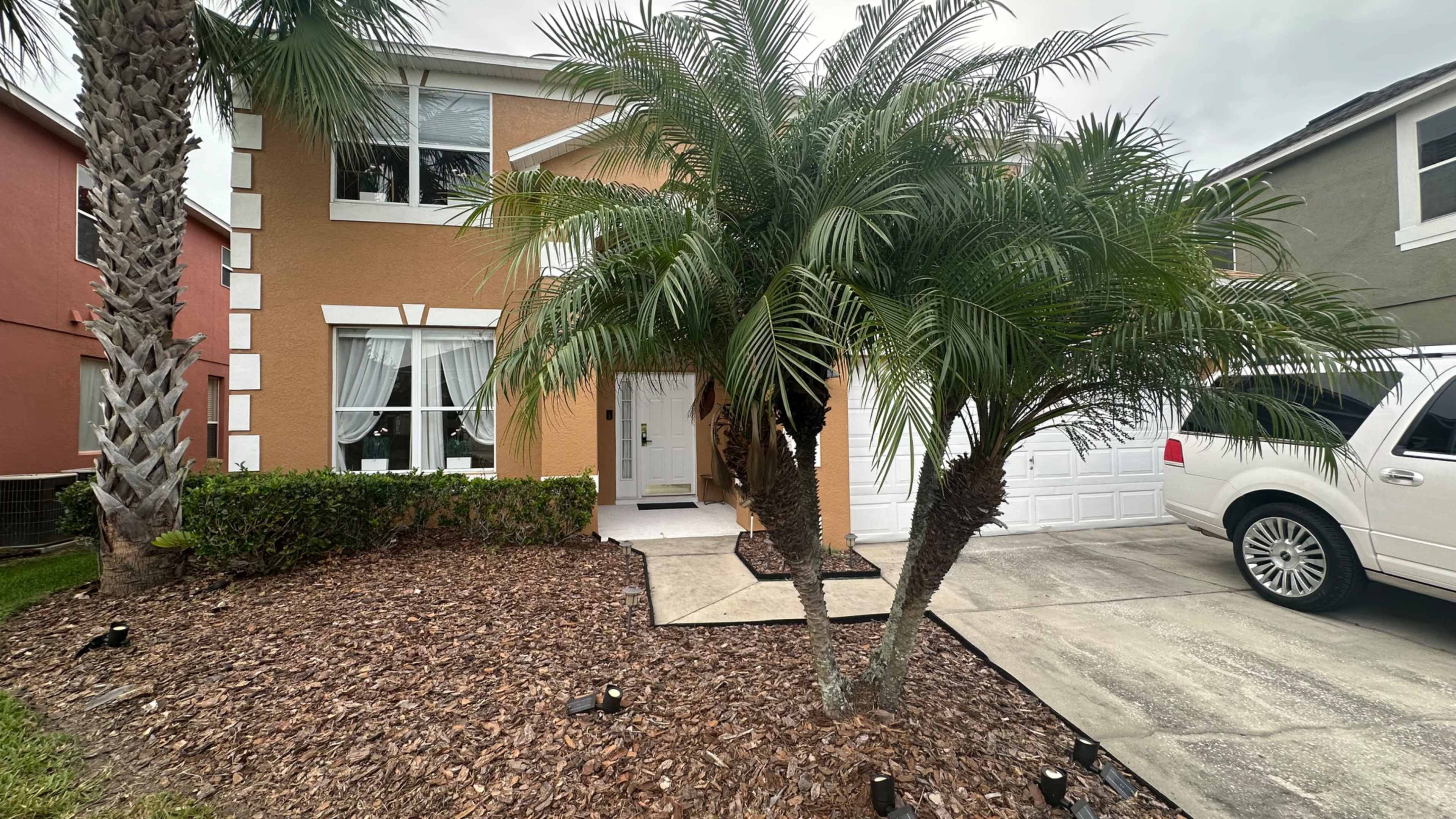 The image shows a two-story house with orange and white exterior walls, featuring palm trees and landscaping in the front yard, along with a parked white vehicle in the driveway.