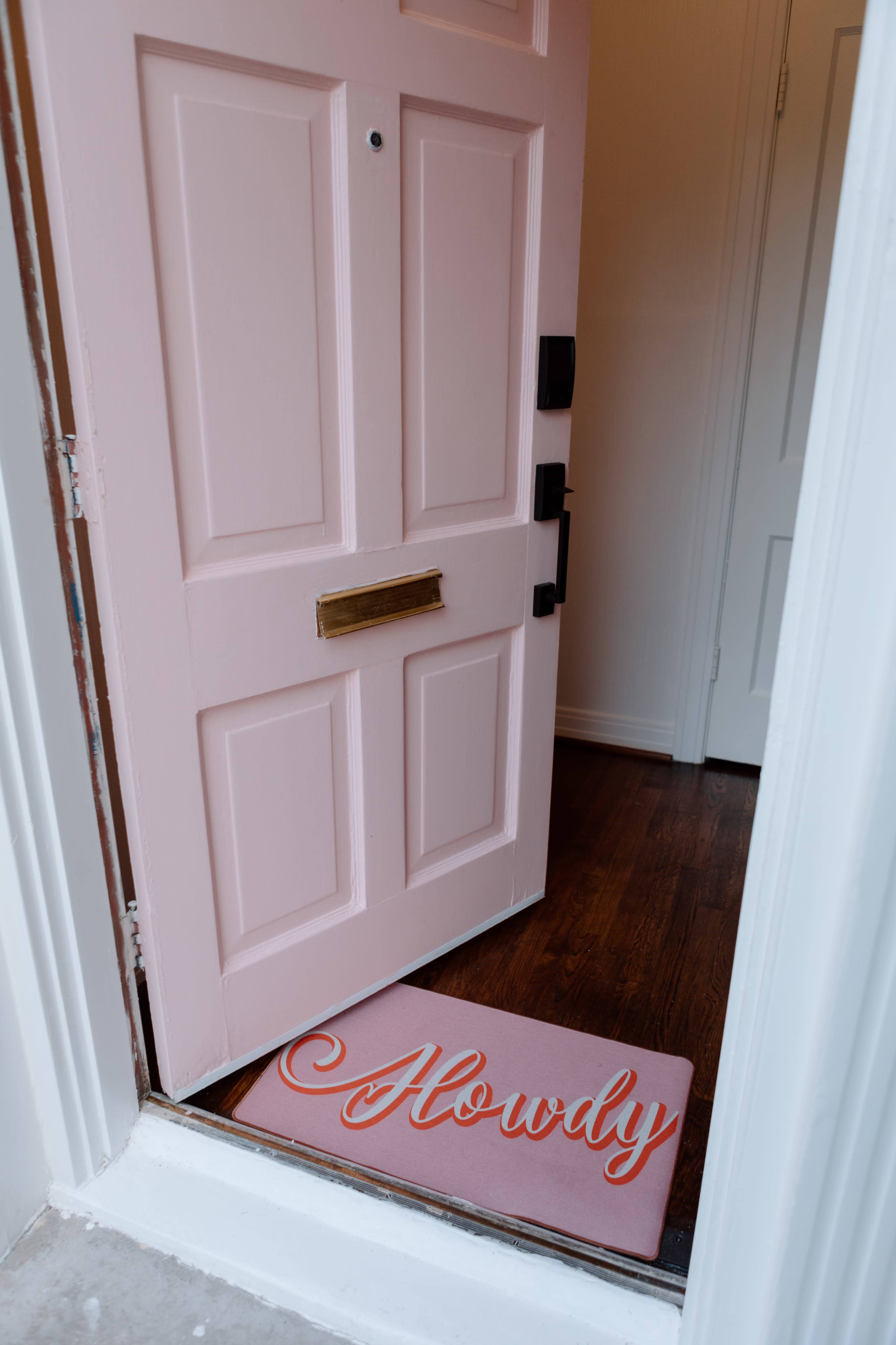 A light pink door stands open, revealing a welcome mat with the word "Howdy" on it.