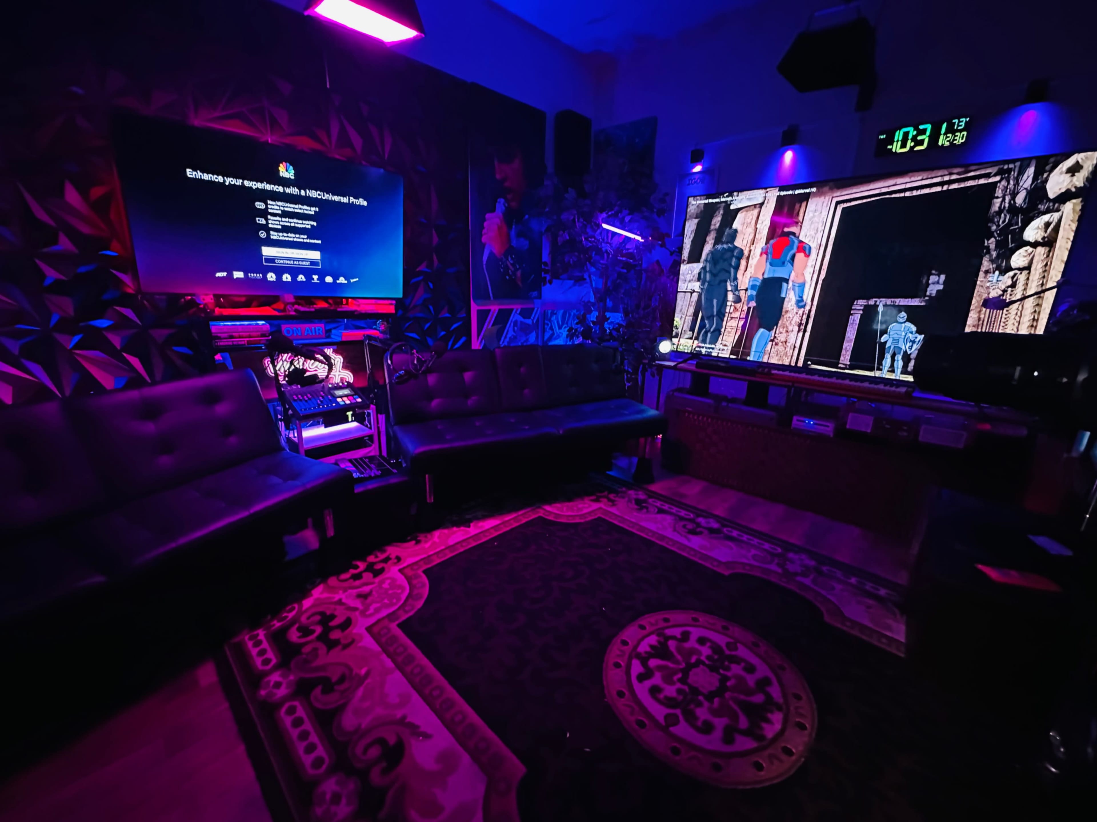 The image shows a dimly lit room with two large screens displaying various content, surrounded by modern seating and colorful lighting effects.