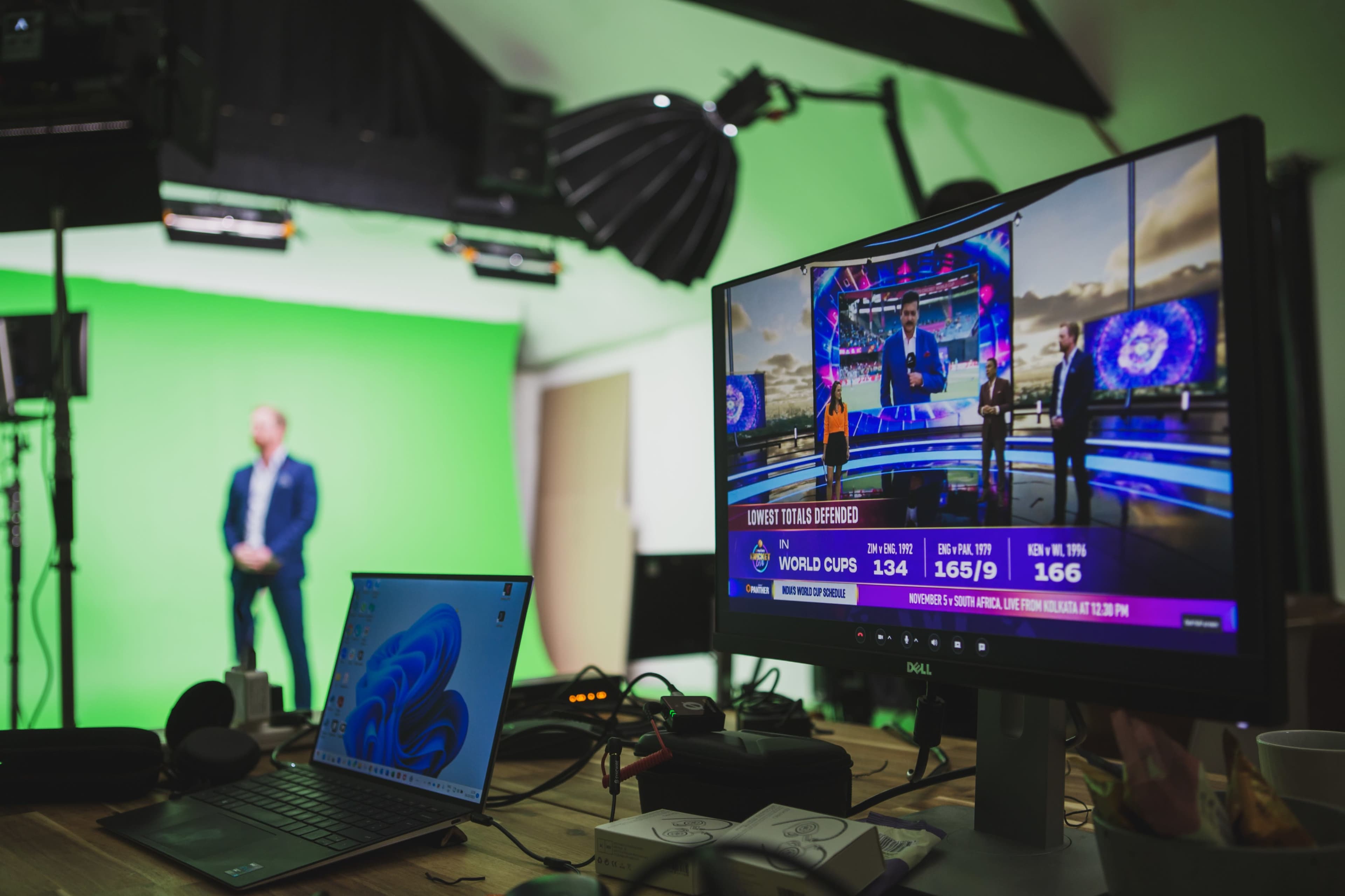 A computer monitor displaying a sports news broadcast is positioned on a desk with various equipment, while a presenter stands in front of a green screen in the background.
