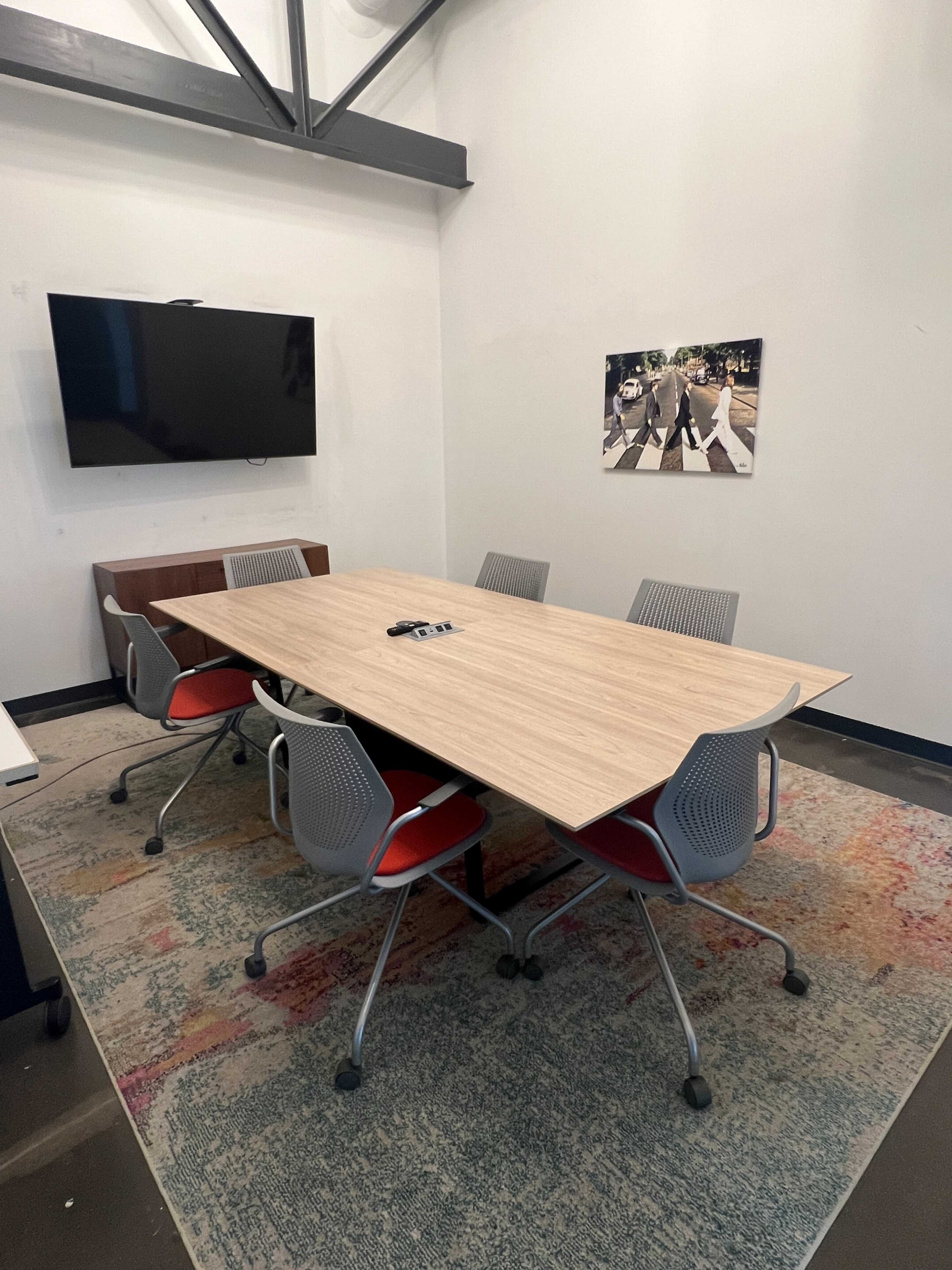 The image shows a conference room with a wooden table surrounded by five chairs, a television mounted on the wall, and a colorful rug on the floor.