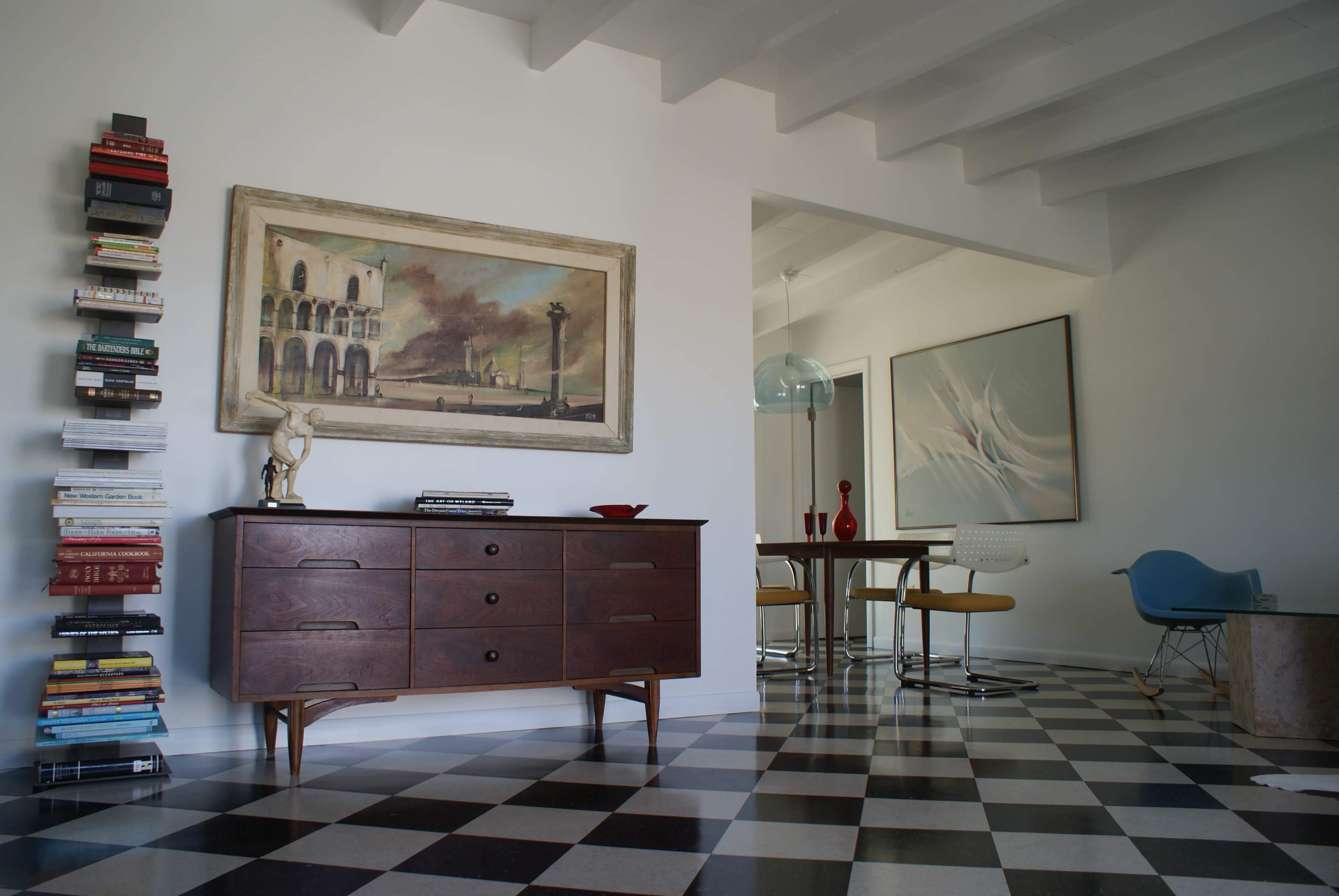 A modern interior featuring a checkered floor, a wooden sideboard with books and decorative items, and a dining area with a round table and mid-century chairs.