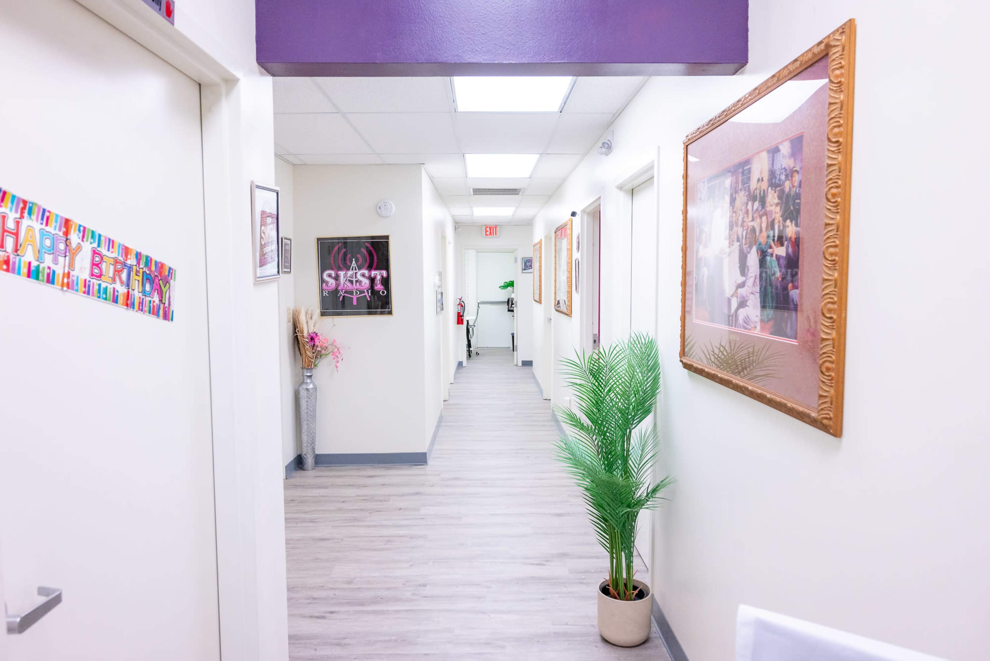 A hallway with light-colored walls, framed pictures, and a potted plant along the right side.