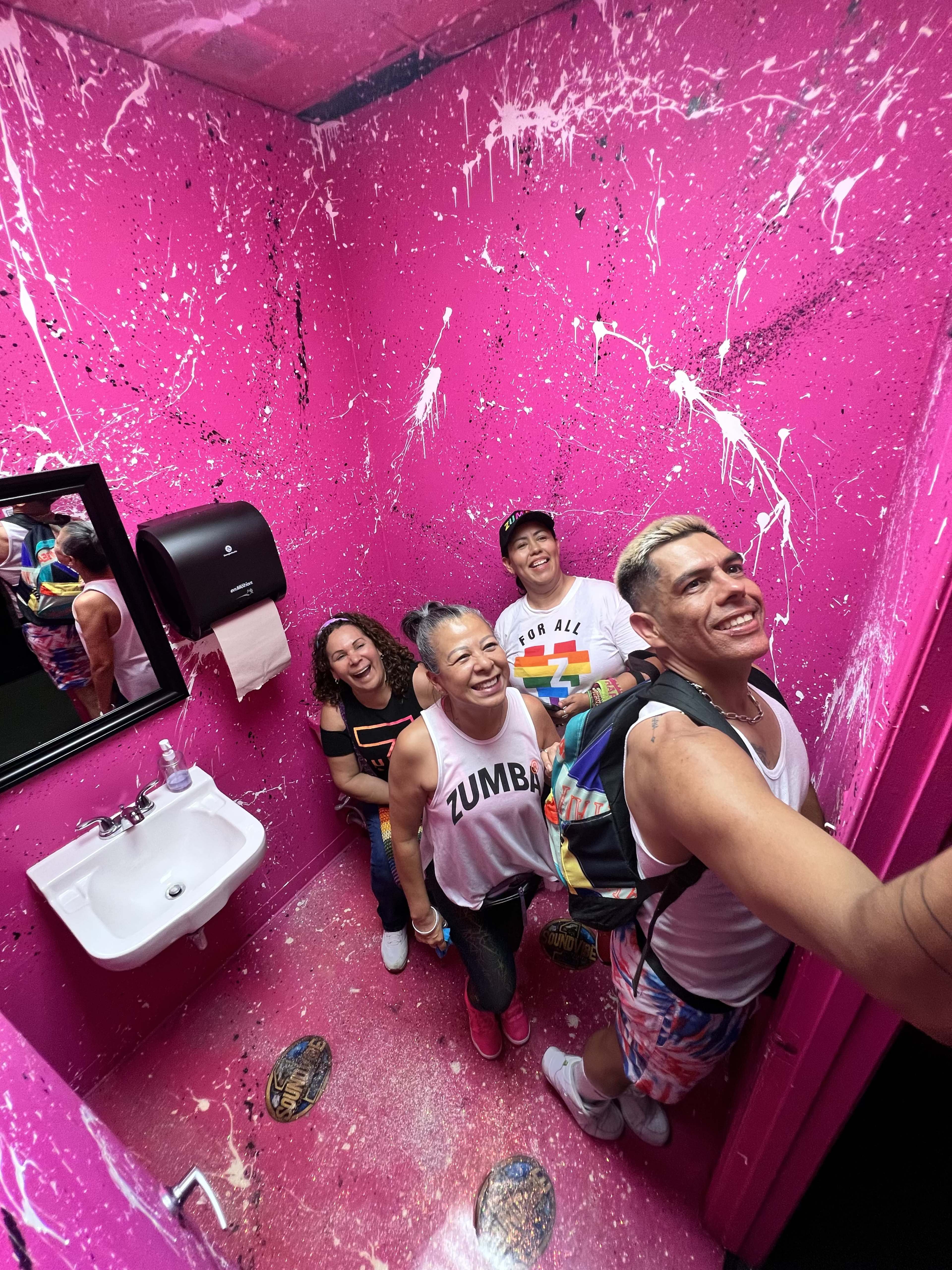 A group of four people is inside a brightly painted pink bathroom with a sink and a mirror.