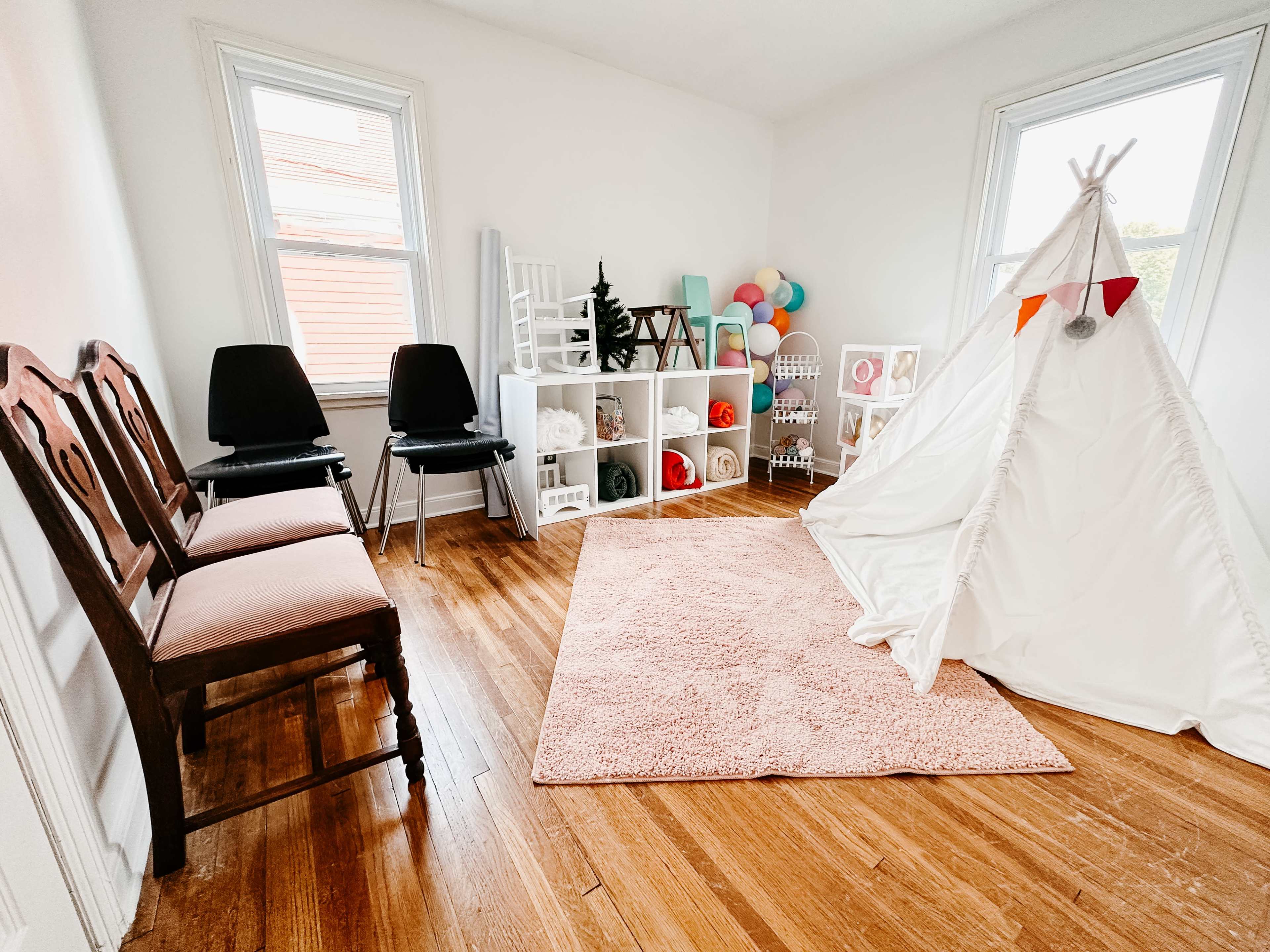 The image shows a bright room with two chairs, a white play tent, a soft pink rug, and storage shelves filled with colorful toys and decorations.