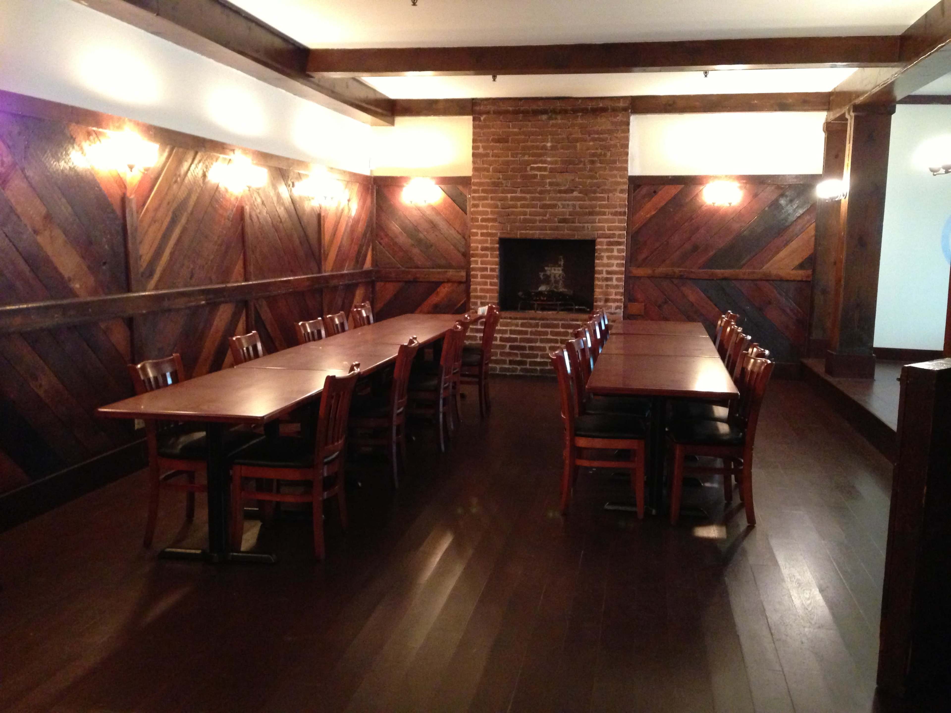 A dining room with two long wooden tables arranged in the center, flanked by wooden chairs, and a brick fireplace on one wall.