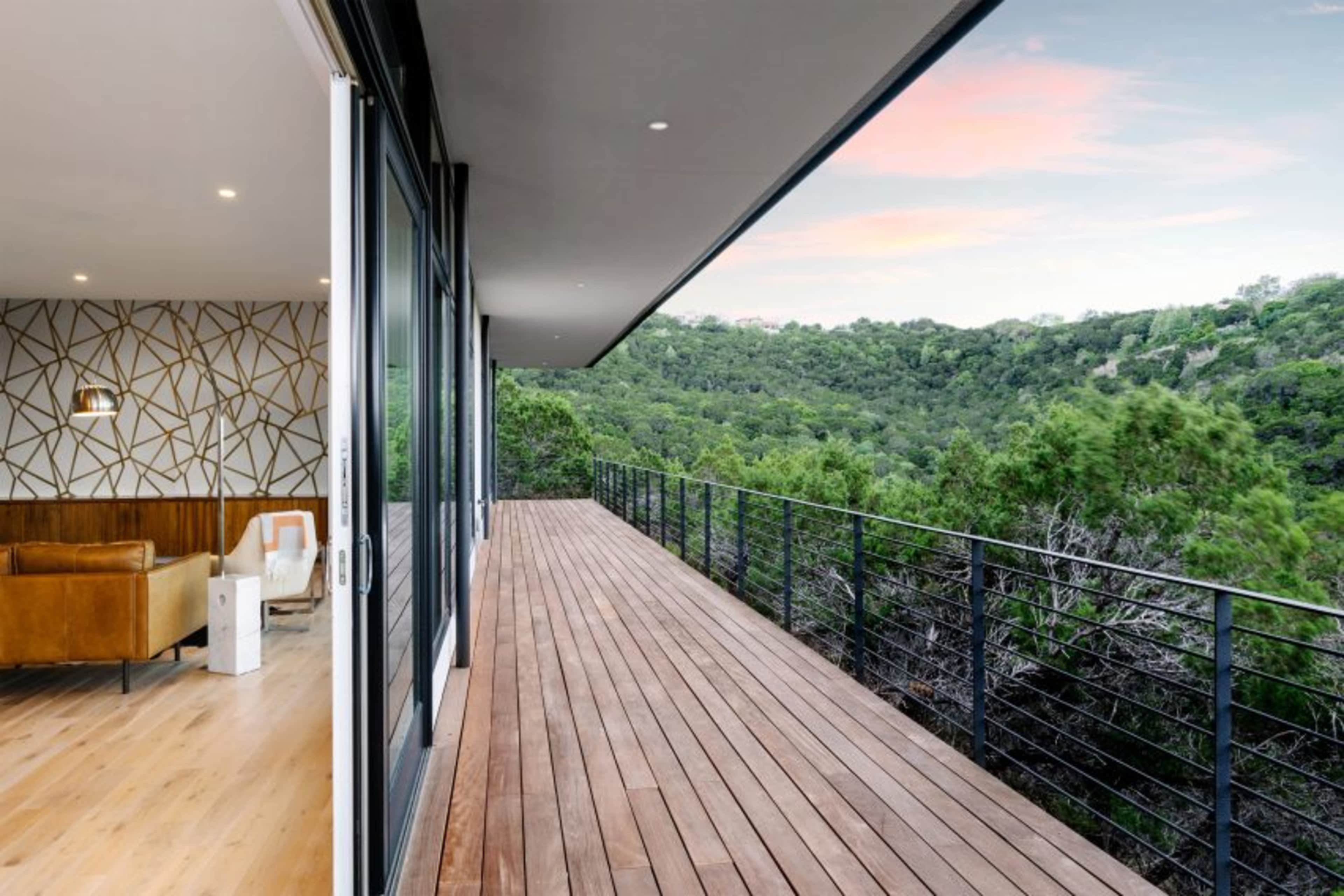 The image shows a wooden deck extending from a modern interior space, overlooking a green hillside under a colorful sky.