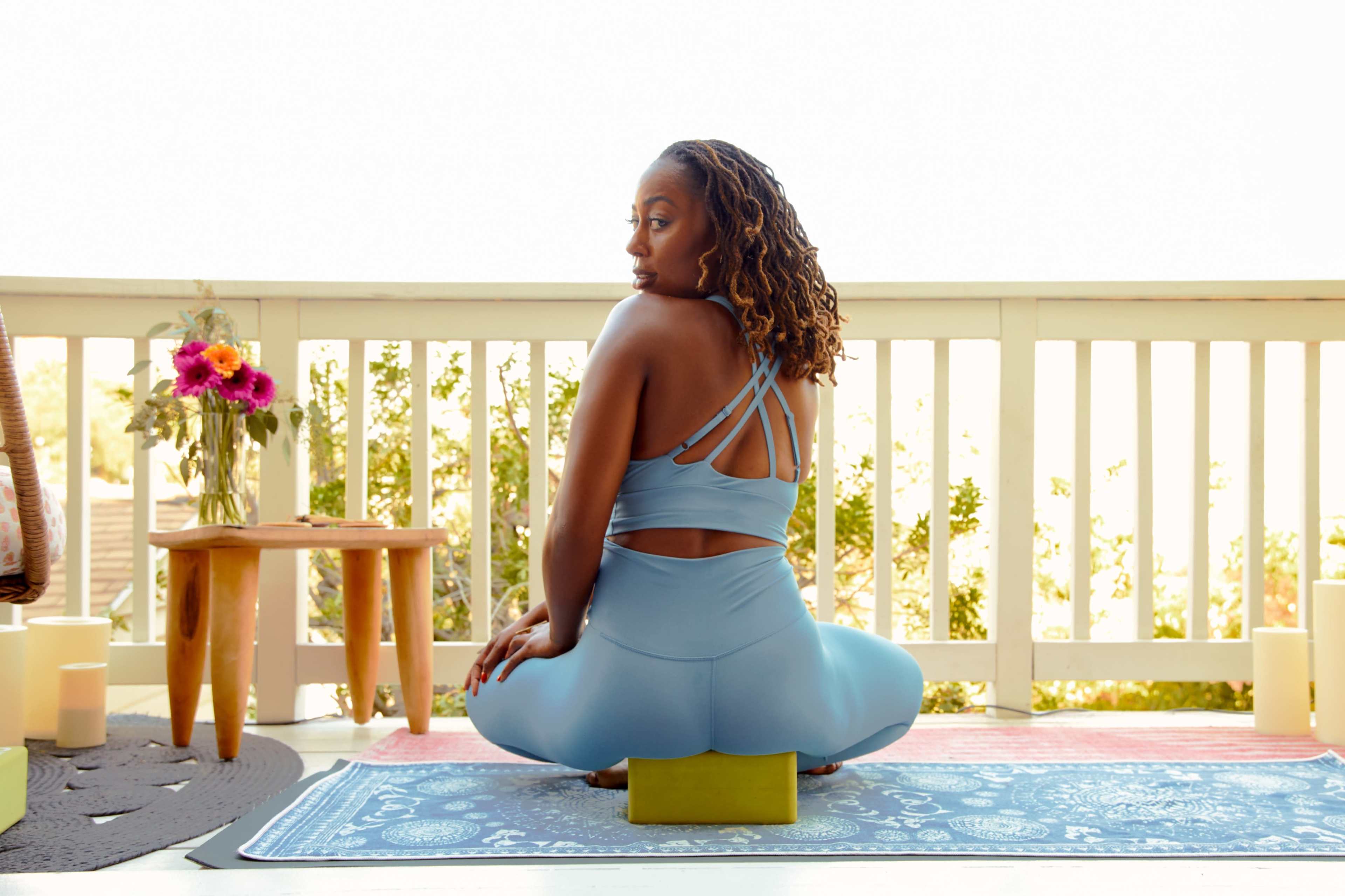 A woman in a blue yoga outfit sits on a mat in a peaceful outdoor setting, with plants and candles surrounding her.