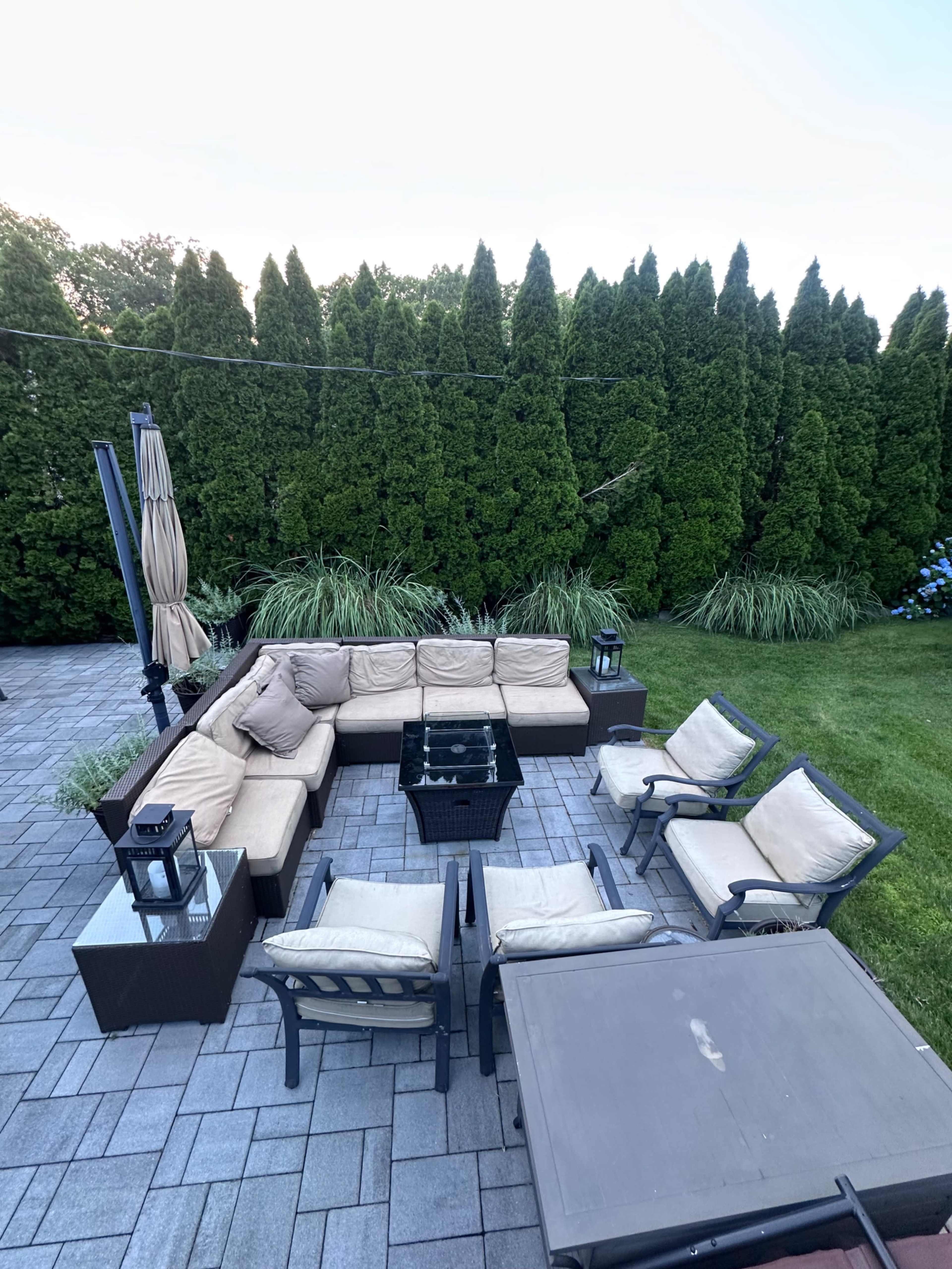 The image shows a landscaped patio area with an outdoor seating arrangement consisting of a sectional sofa and several chairs around a central table, surrounded by tall green bushes.
