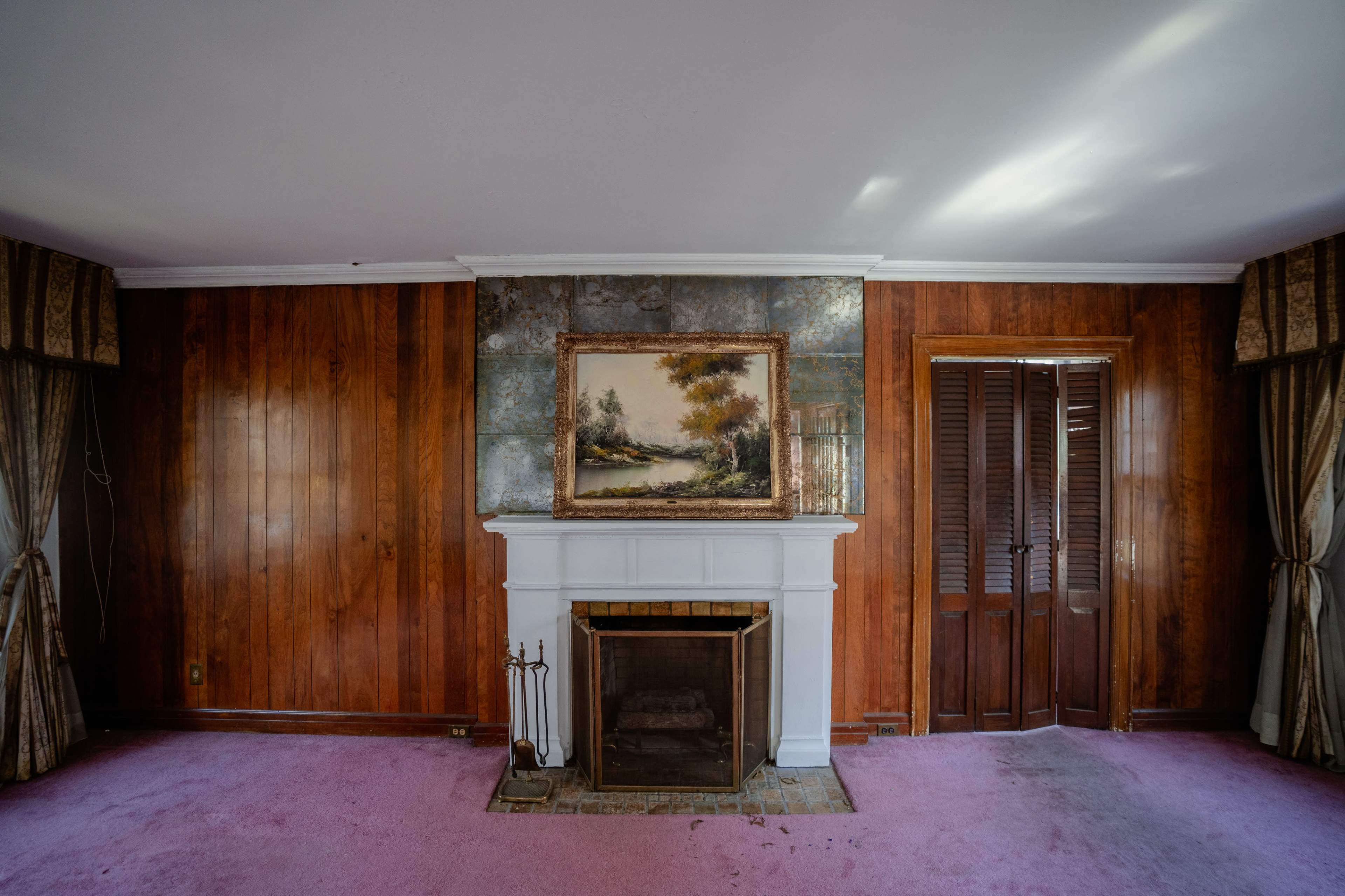 The image shows a room with wooden paneling, a white fireplace, and a painting of a landscape hanging above it, along with pink carpet and closed wooden shutters.