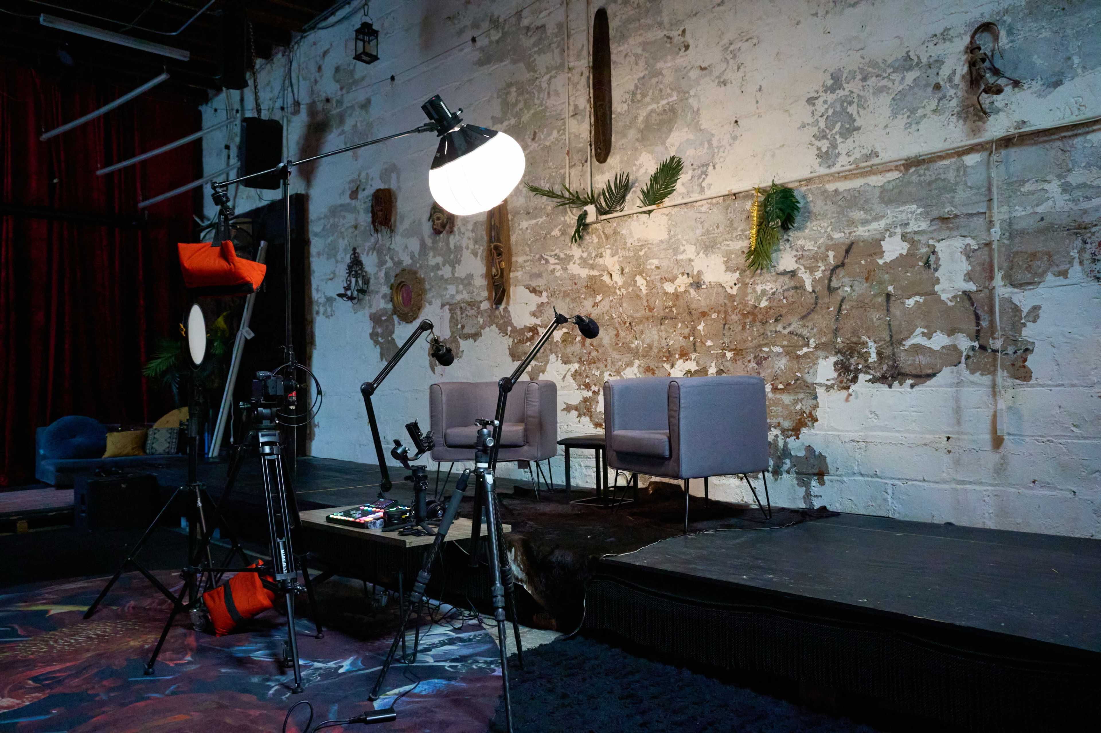 The image shows a staged interview setup with two gray chairs on a raised platform, surrounded by various lighting equipment and a textured wall with decorative elements.