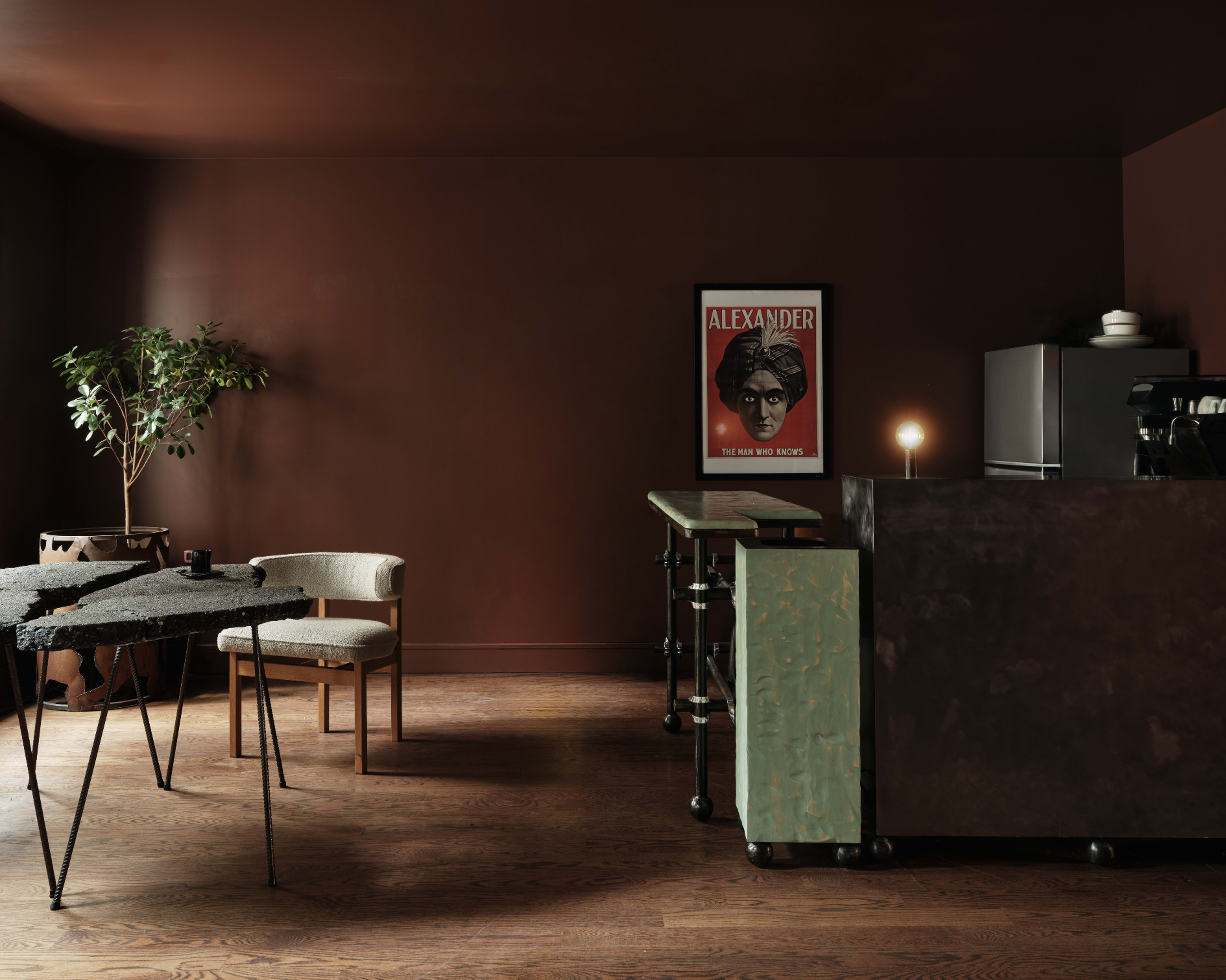The image shows a modern interior space featuring a dark brown wall, a framed poster, a small table with two chairs, and a counter with a coffee machine.