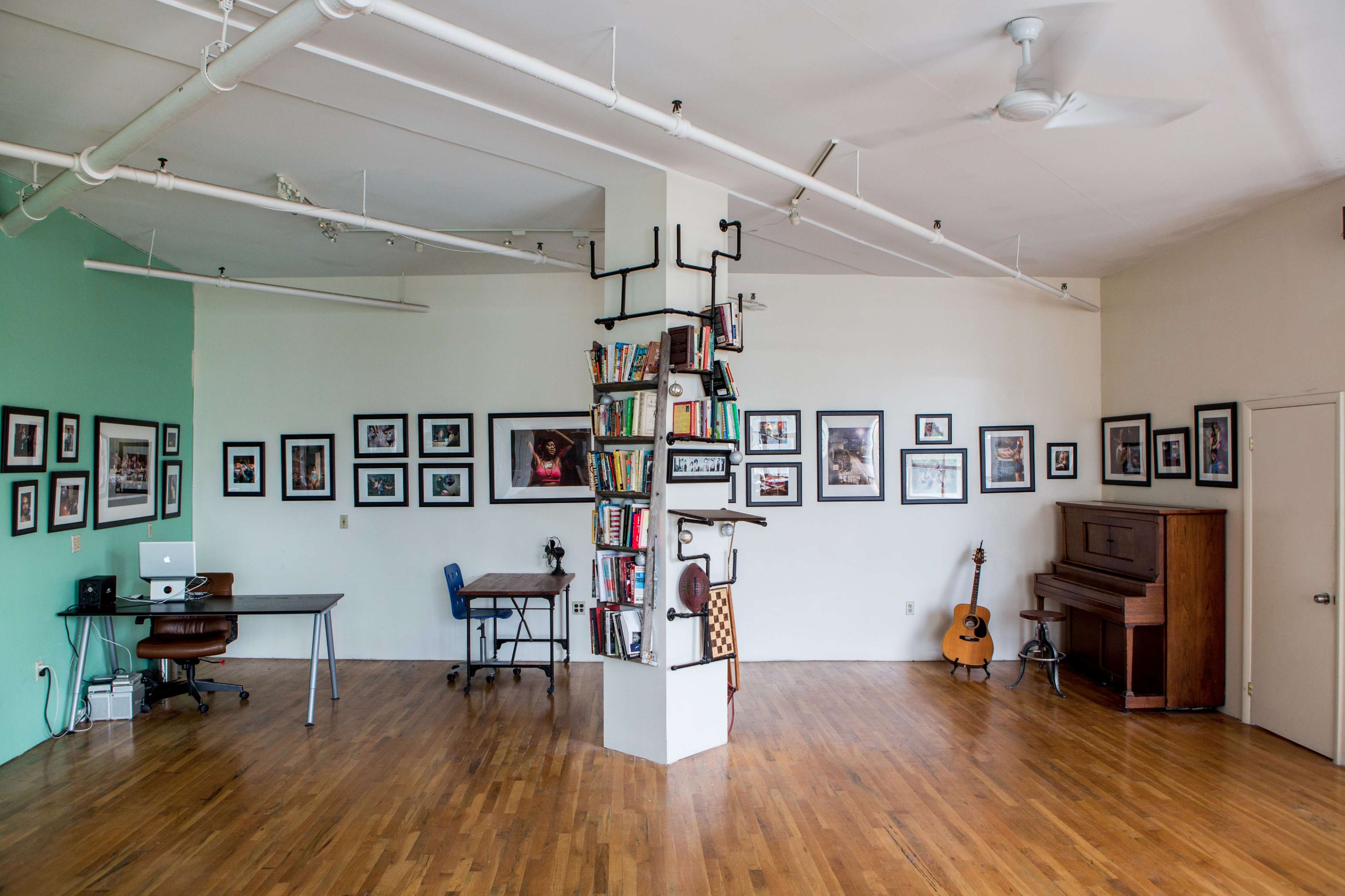 A spacious room with wooden flooring, featuring a work desk, a piano, bookshelves built around a central pillar, and framed photographs on the walls.