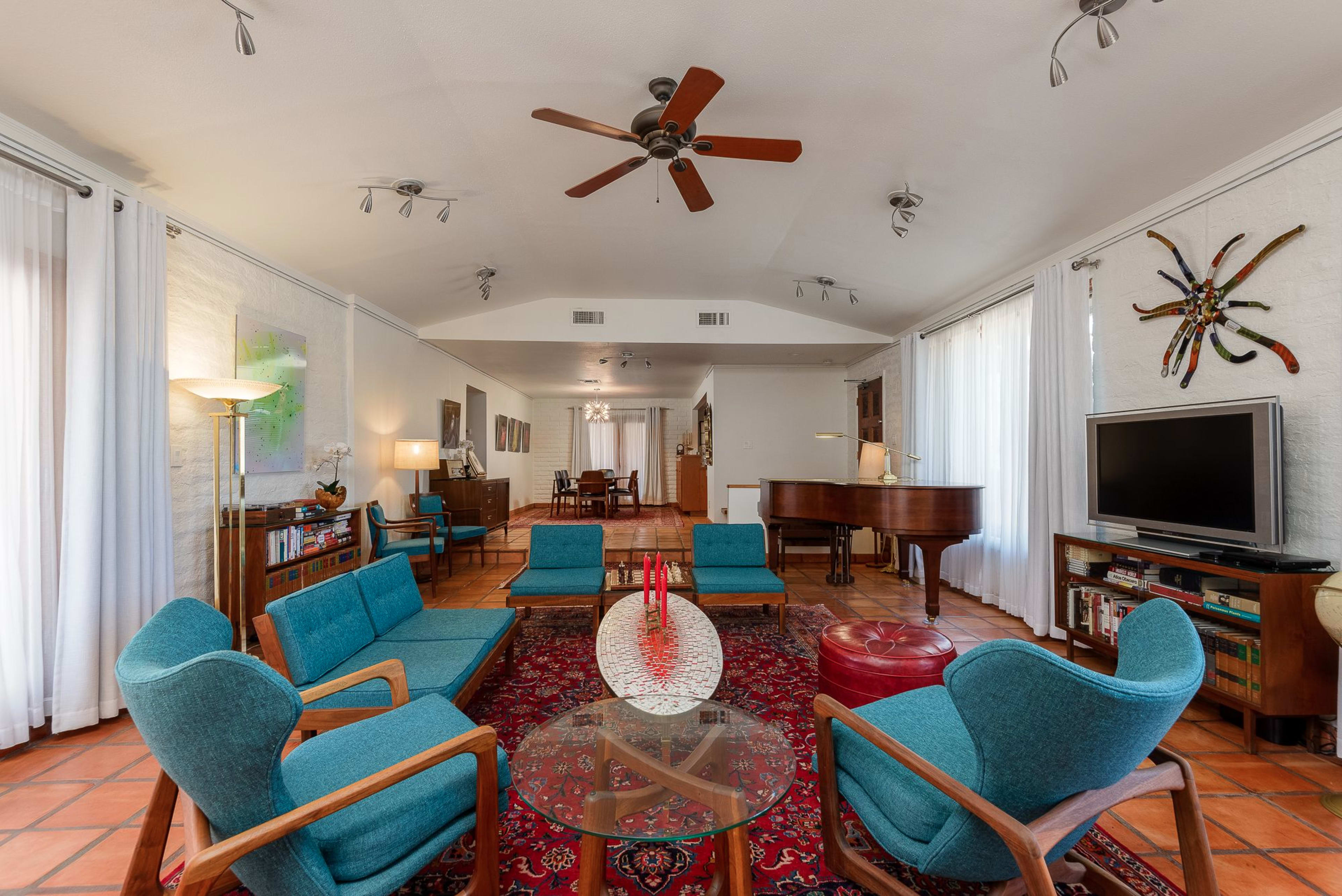 A spacious living room with turquoise chairs arranged around a glass coffee table and a patterned rug, complemented by ceiling fans and large windows.