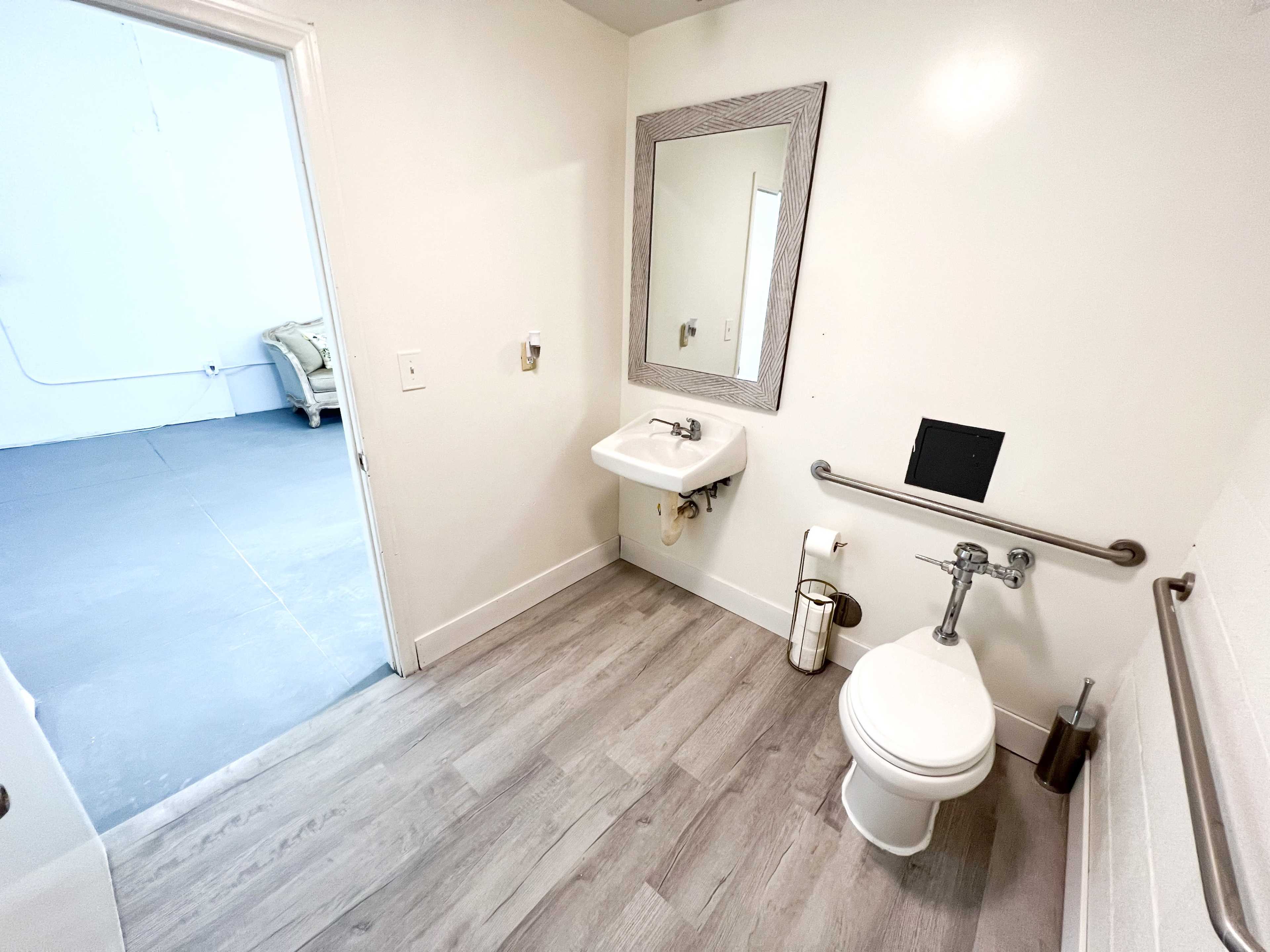 A clean bathroom features a toilet, wall-mounted sink, and a large mirror, with a door leading to an adjacent room.