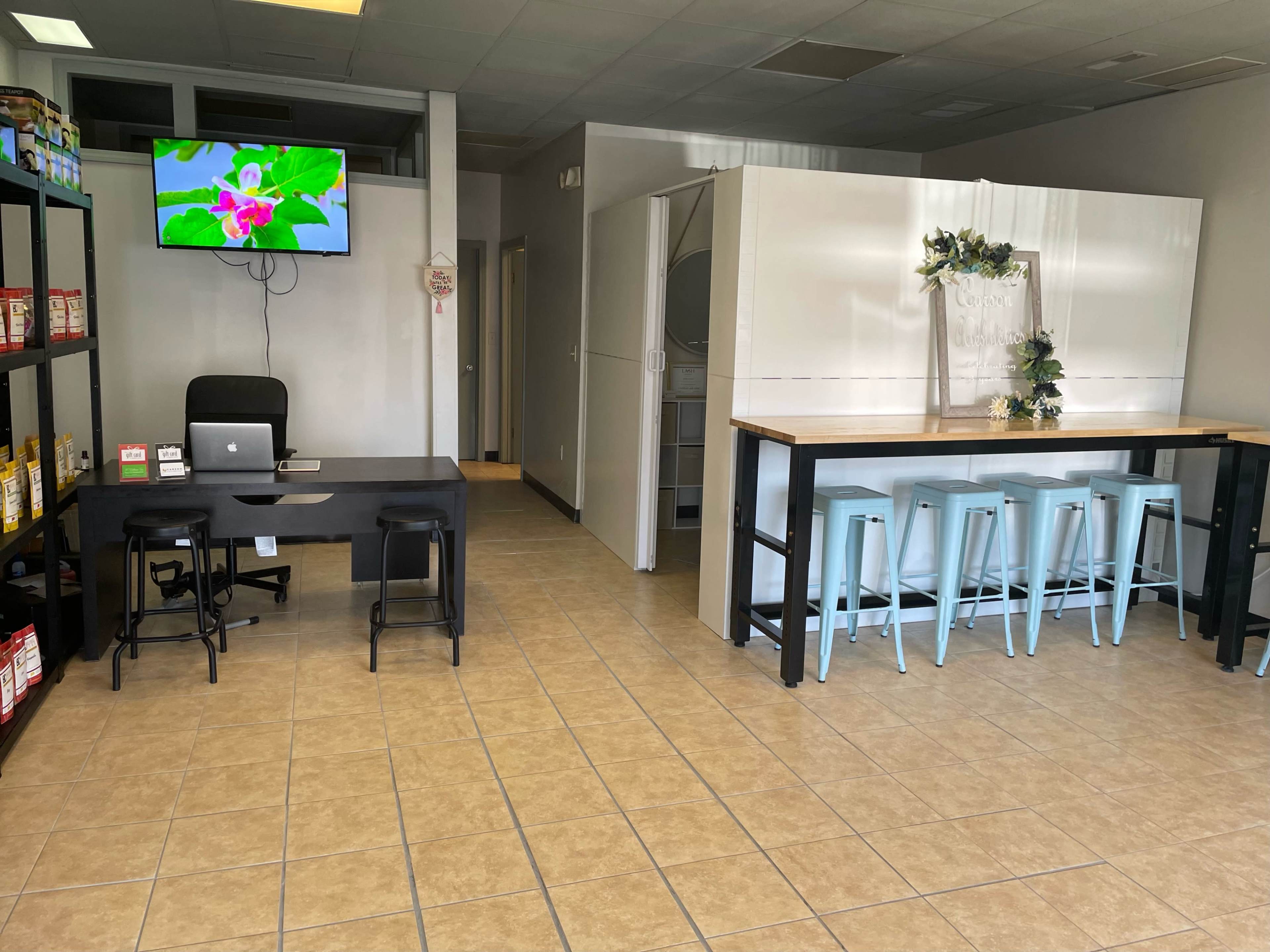 The image shows a spacious interior of a commercial space featuring a reception area with a desk and a TV, alongside a dining area with a long table and stools.