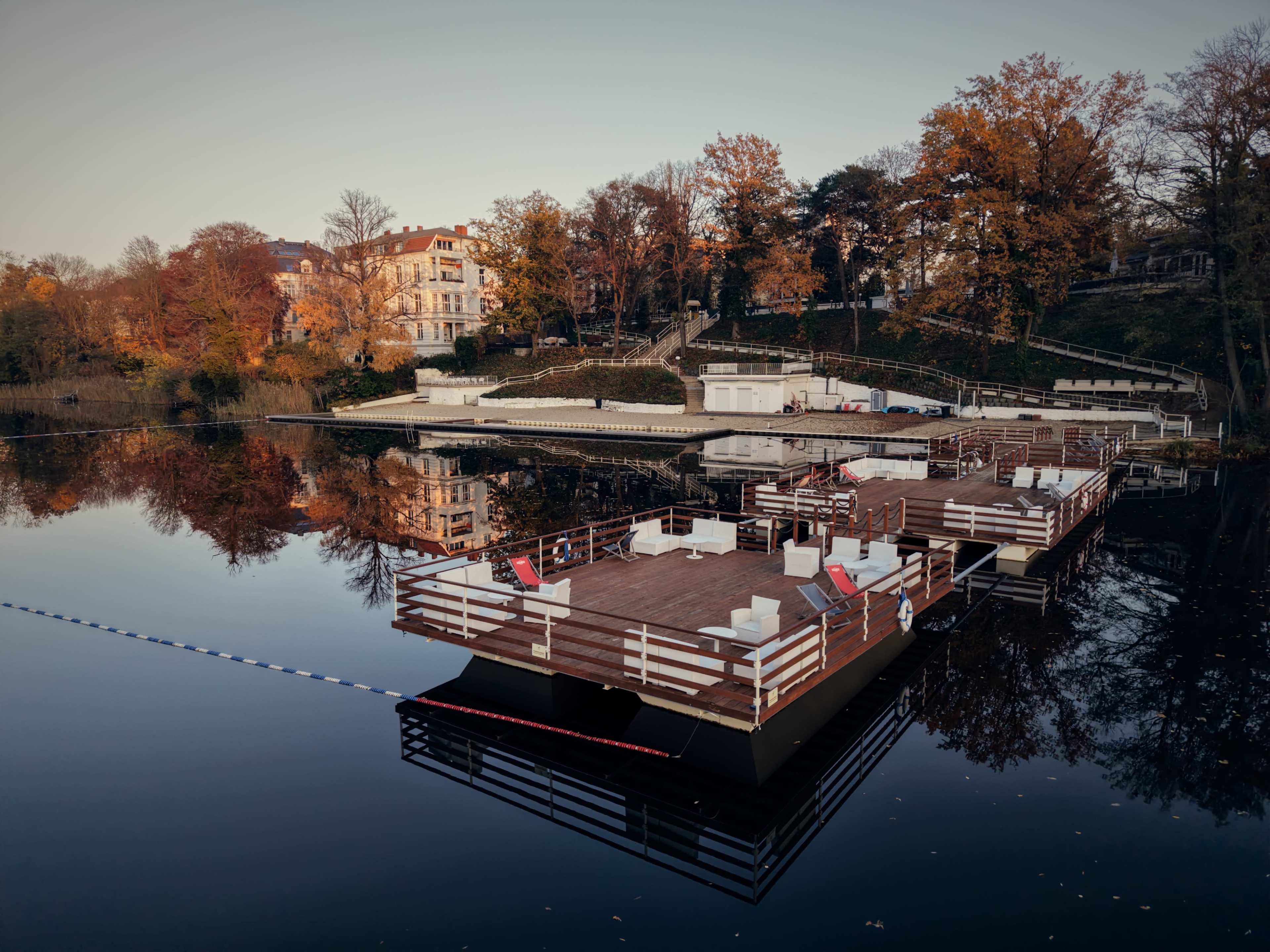 A floating platform with white furniture is anchored on a calm body of water, surrounded by trees with autumn foliage.