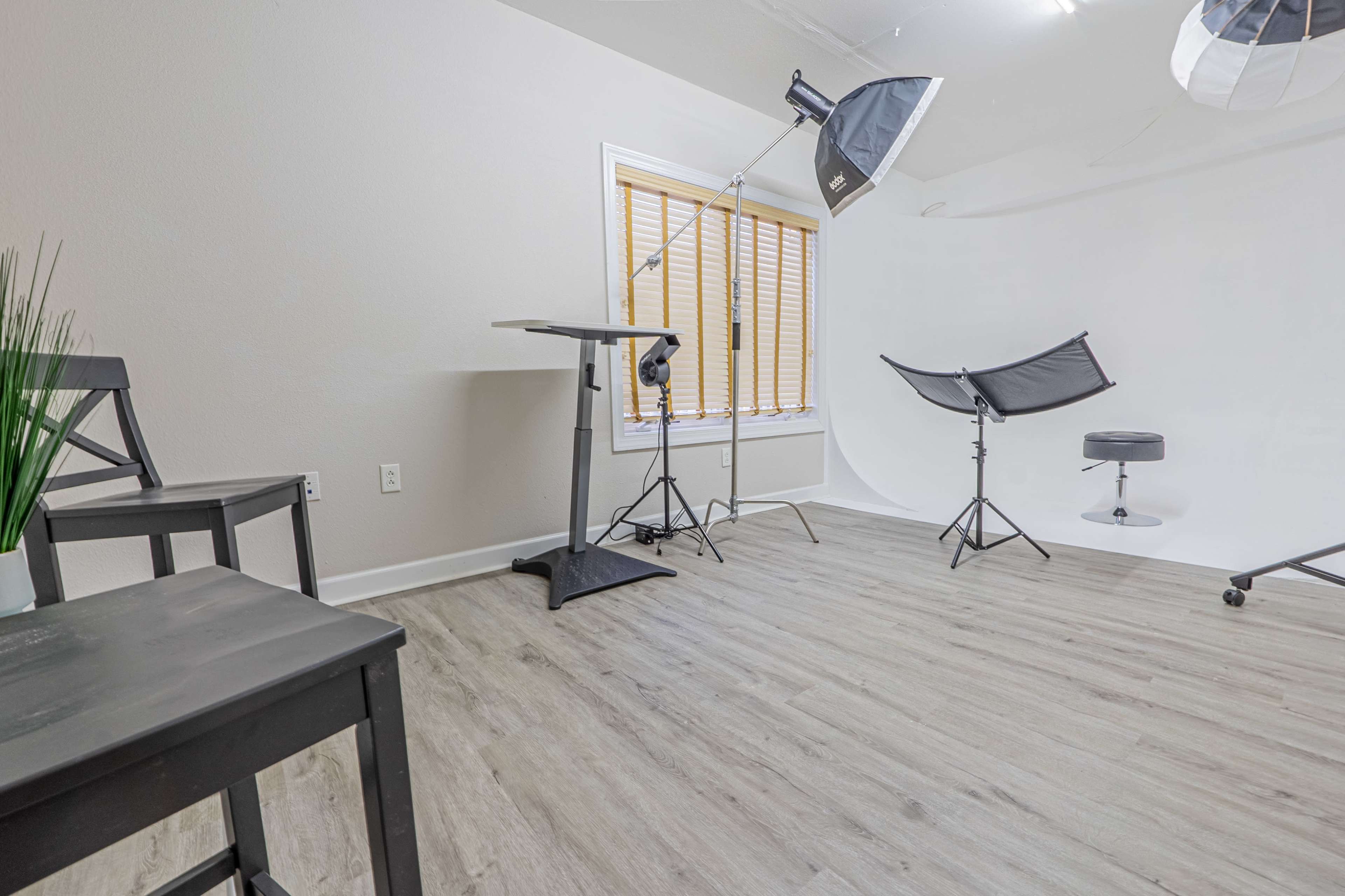 A photography studio with a white backdrop, lighting equipment, and a few chairs arranged near a window.
