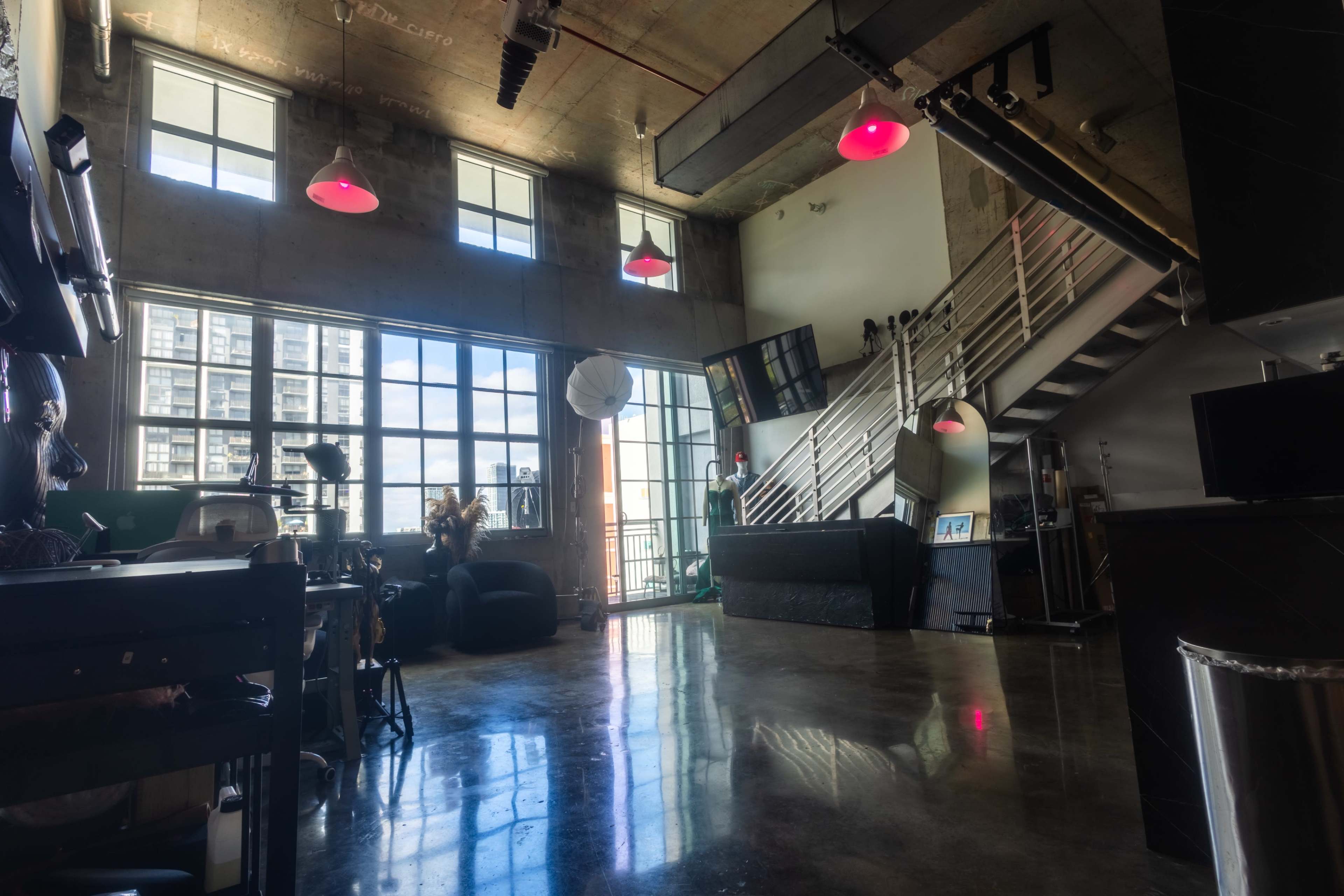 The image shows a spacious, modern loft with large windows, industrial features, and a staircase leading to an upper level.