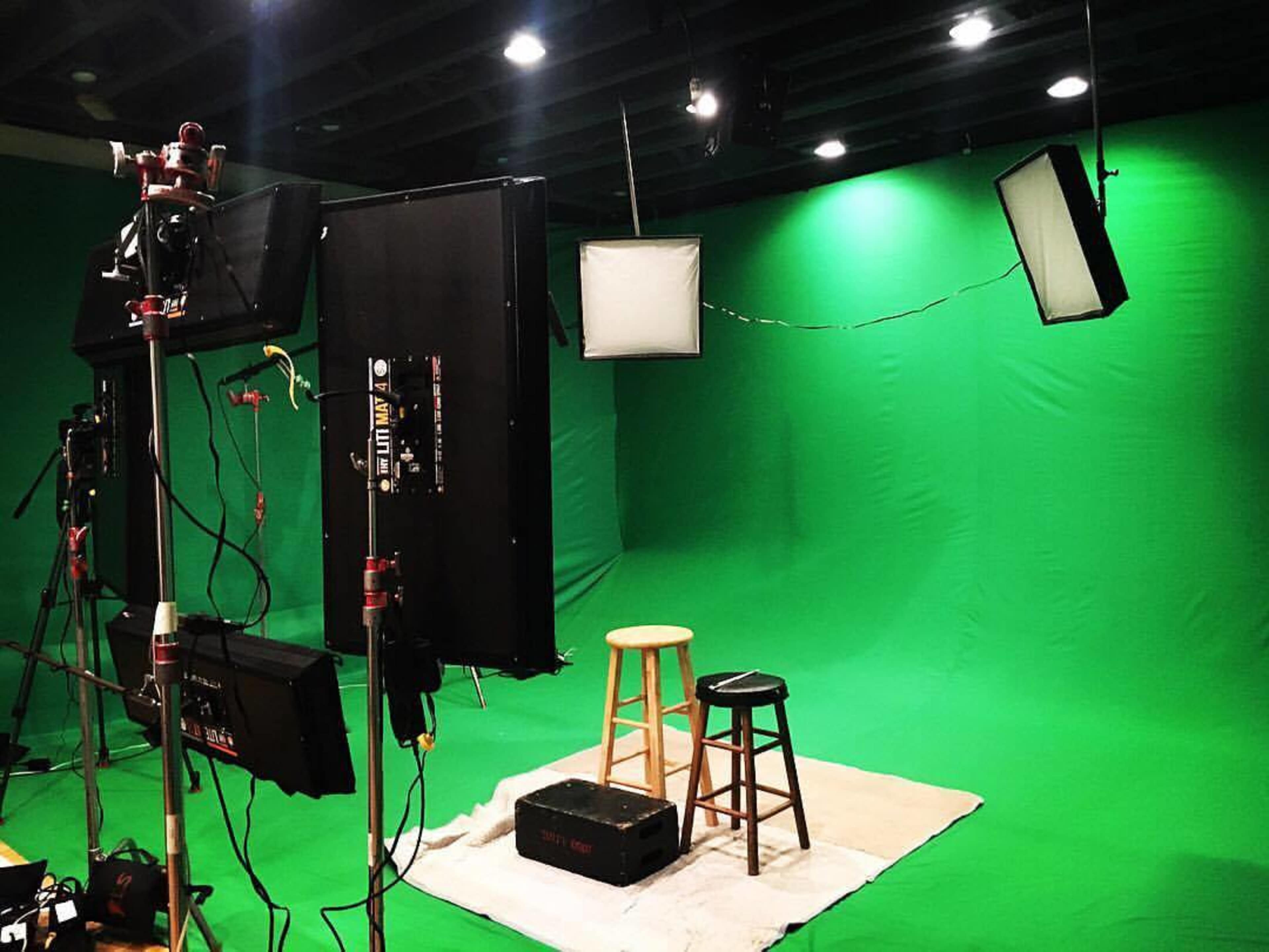 A film set with two wooden stools placed on a light-colored rug in front of a green screen, surrounded by lighting equipment.