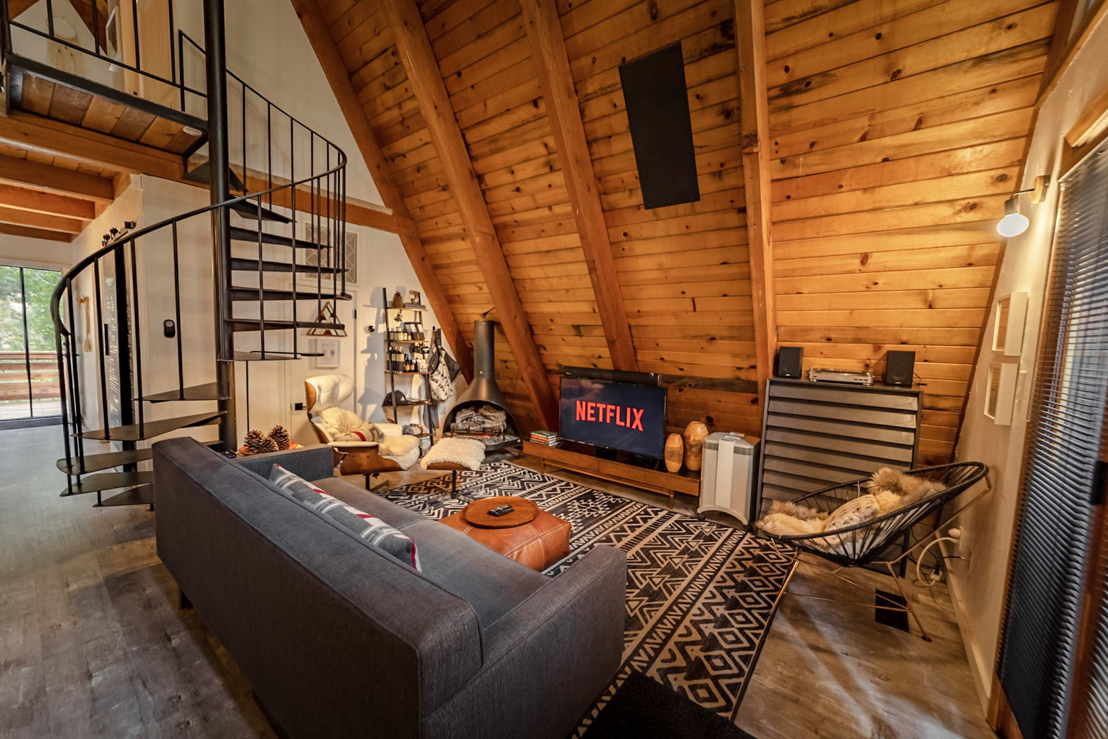 The living room features a high, wooden beamed ceiling, a spiral staircase, a large gray sofa, and a television displaying the Netflix logo.