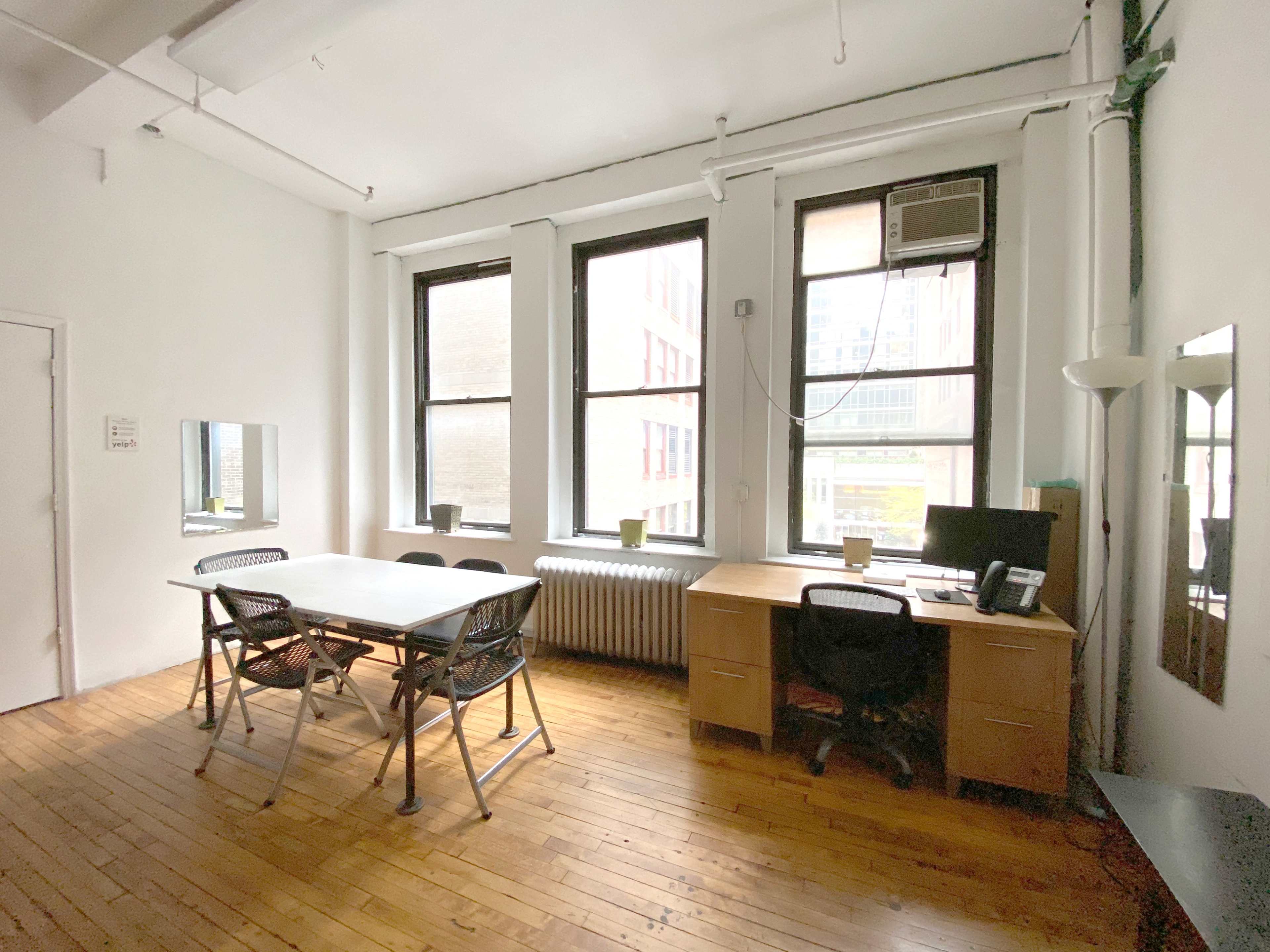 A simple office space with a desk and chairs, featuring large windows, hardwood floors, and a radiator.