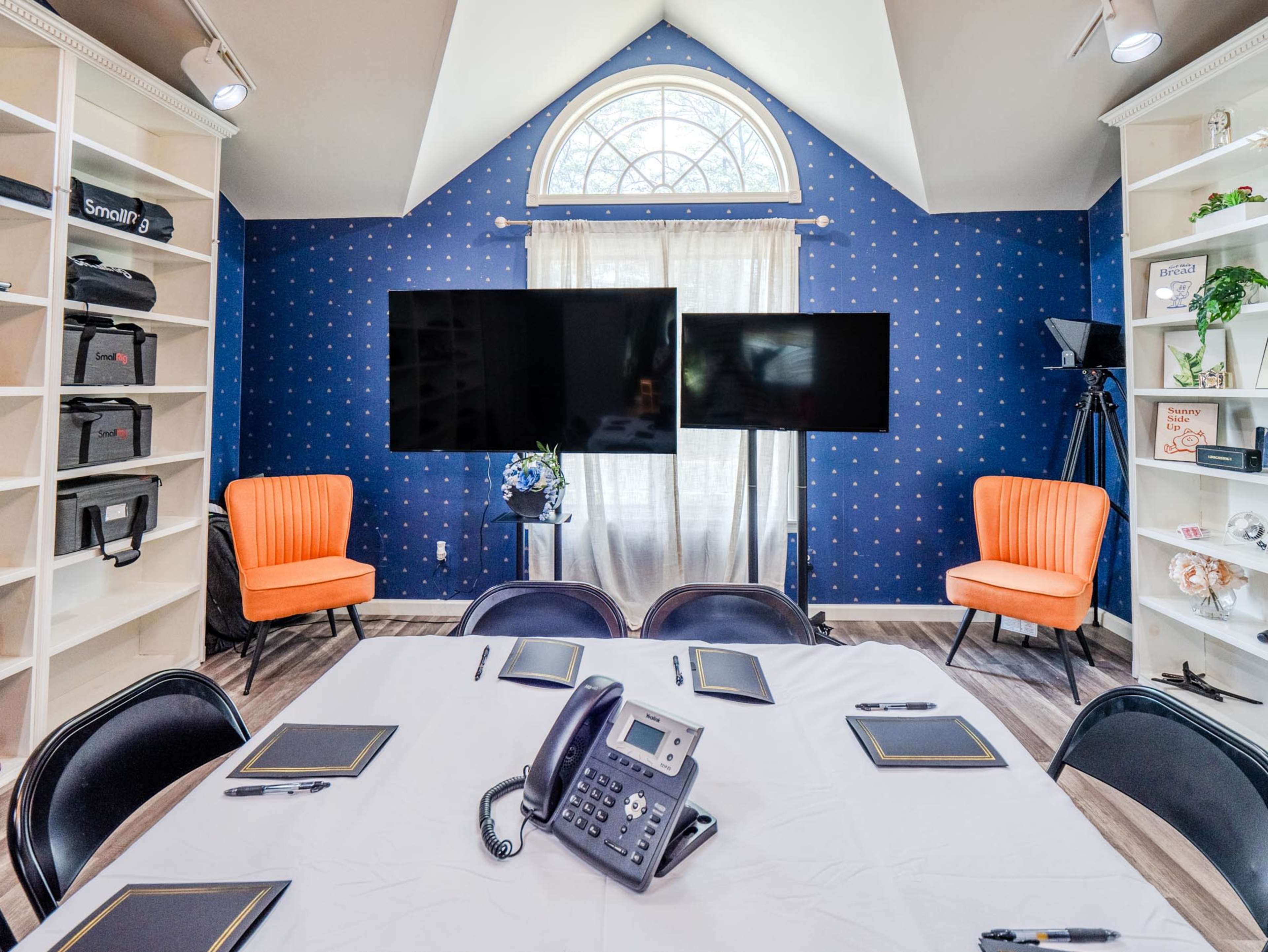 A conference room features a round table with notepads and a phone, flanked by two orange chairs, two television screens, and a large window.