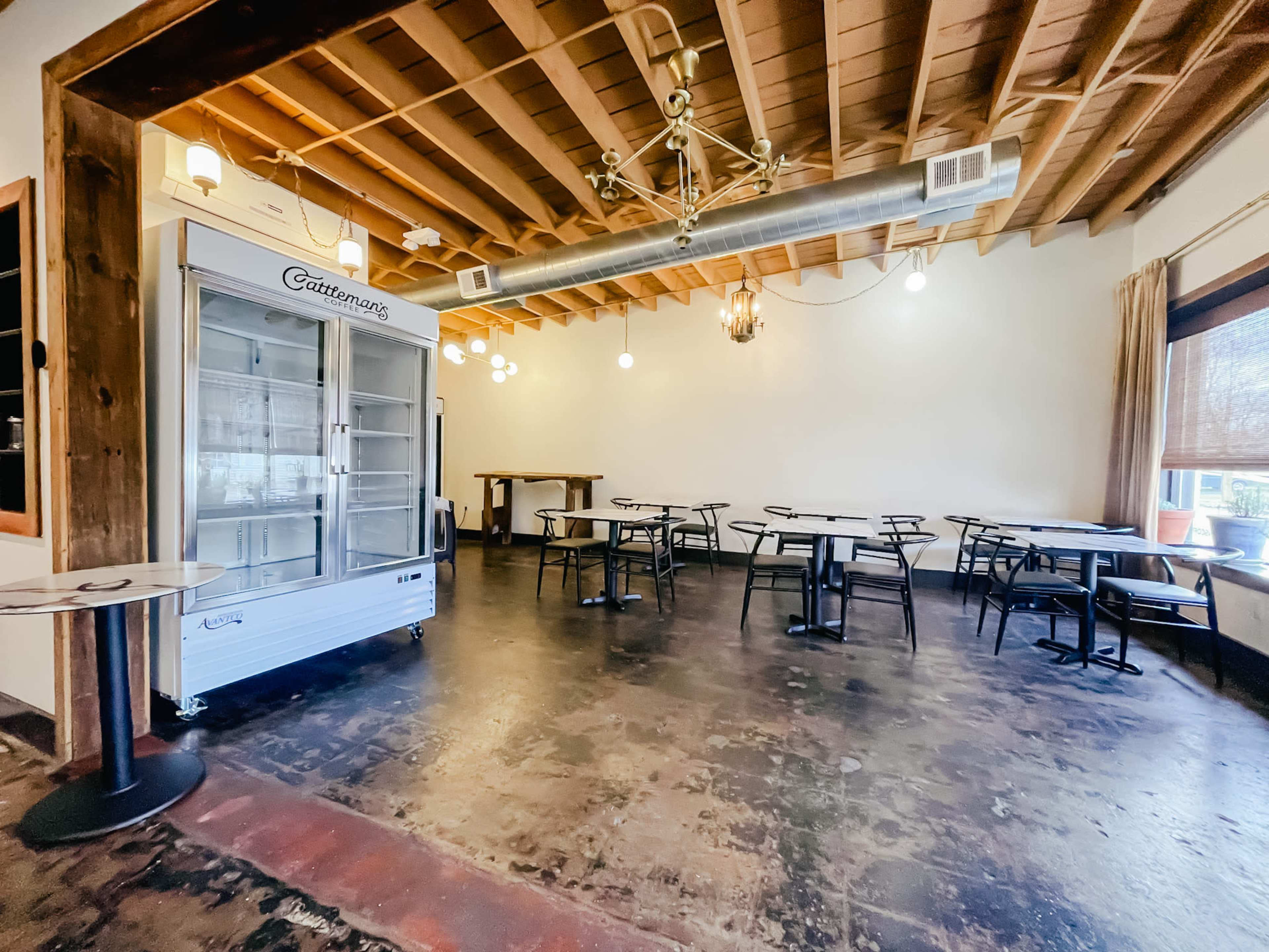 A spacious, empty dining area with wooden ceilings, several tables and chairs, and a glass-fronted refrigerator against one wall.