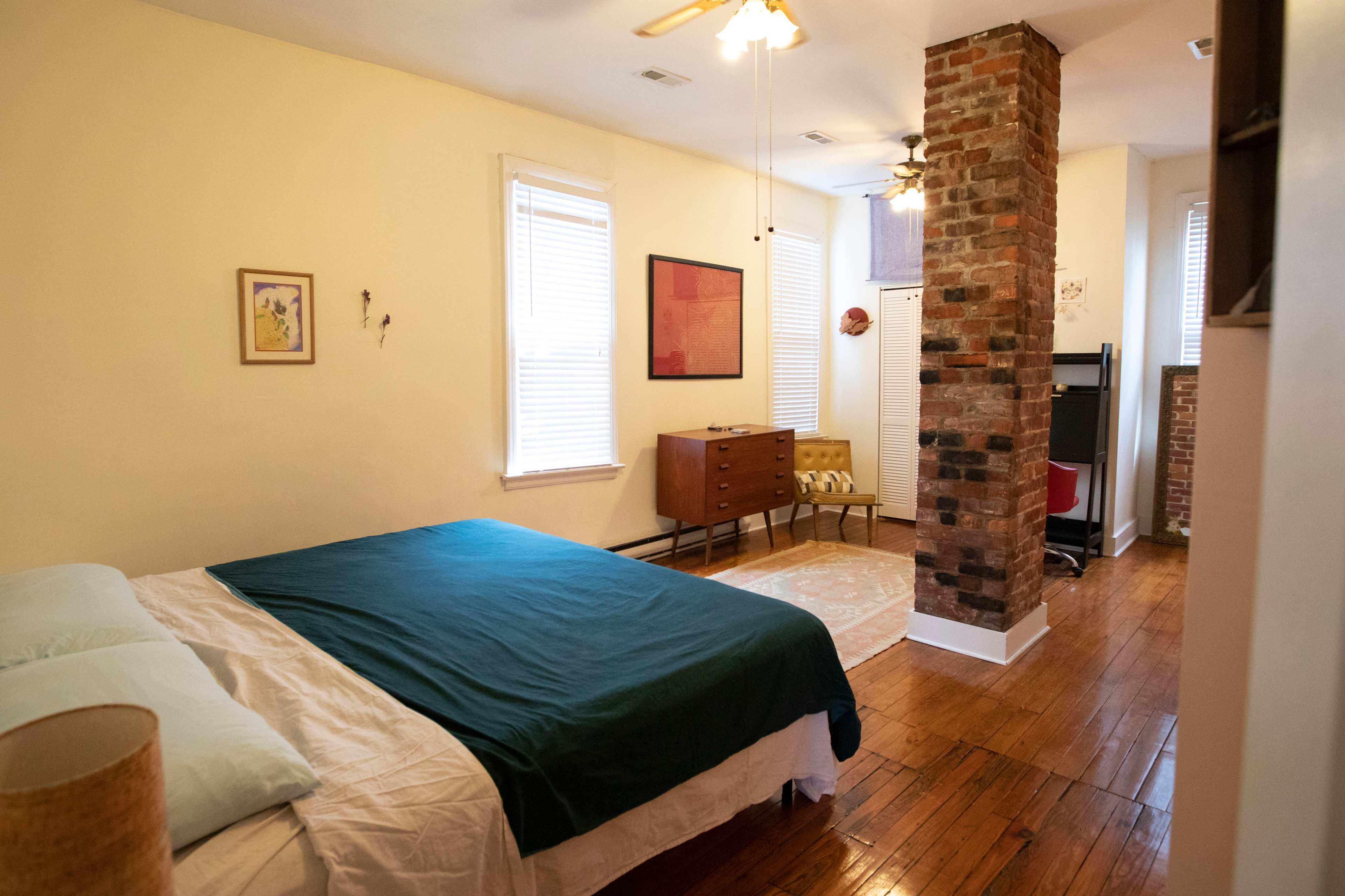 A bedroom features a bed with a green blanket, a wooden dresser, and exposed brick accents.