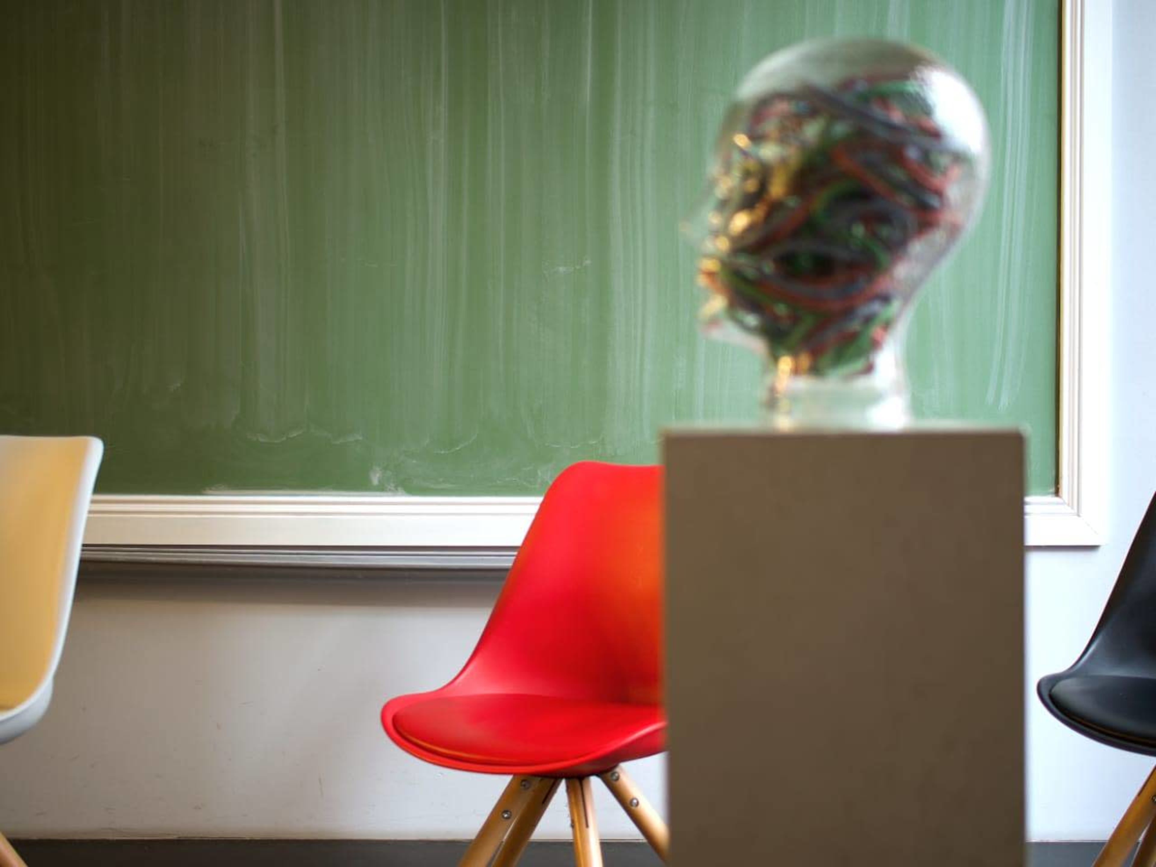 The image shows a red chair and a black chair in front of a green chalkboard, with a transparent head sculpture filled with colorful wires on a pedestal nearby.