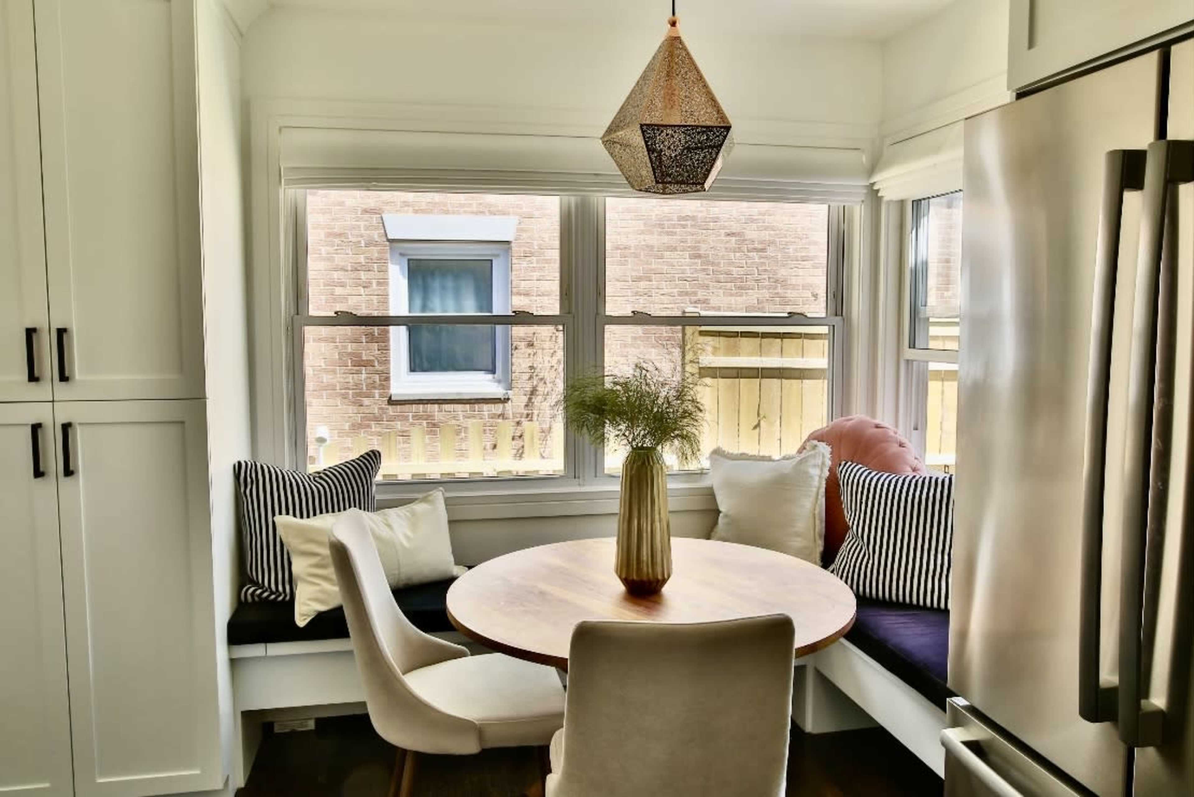 A small dining area features a round table surrounded by a corner bench and chairs, with large windows letting in natural light.