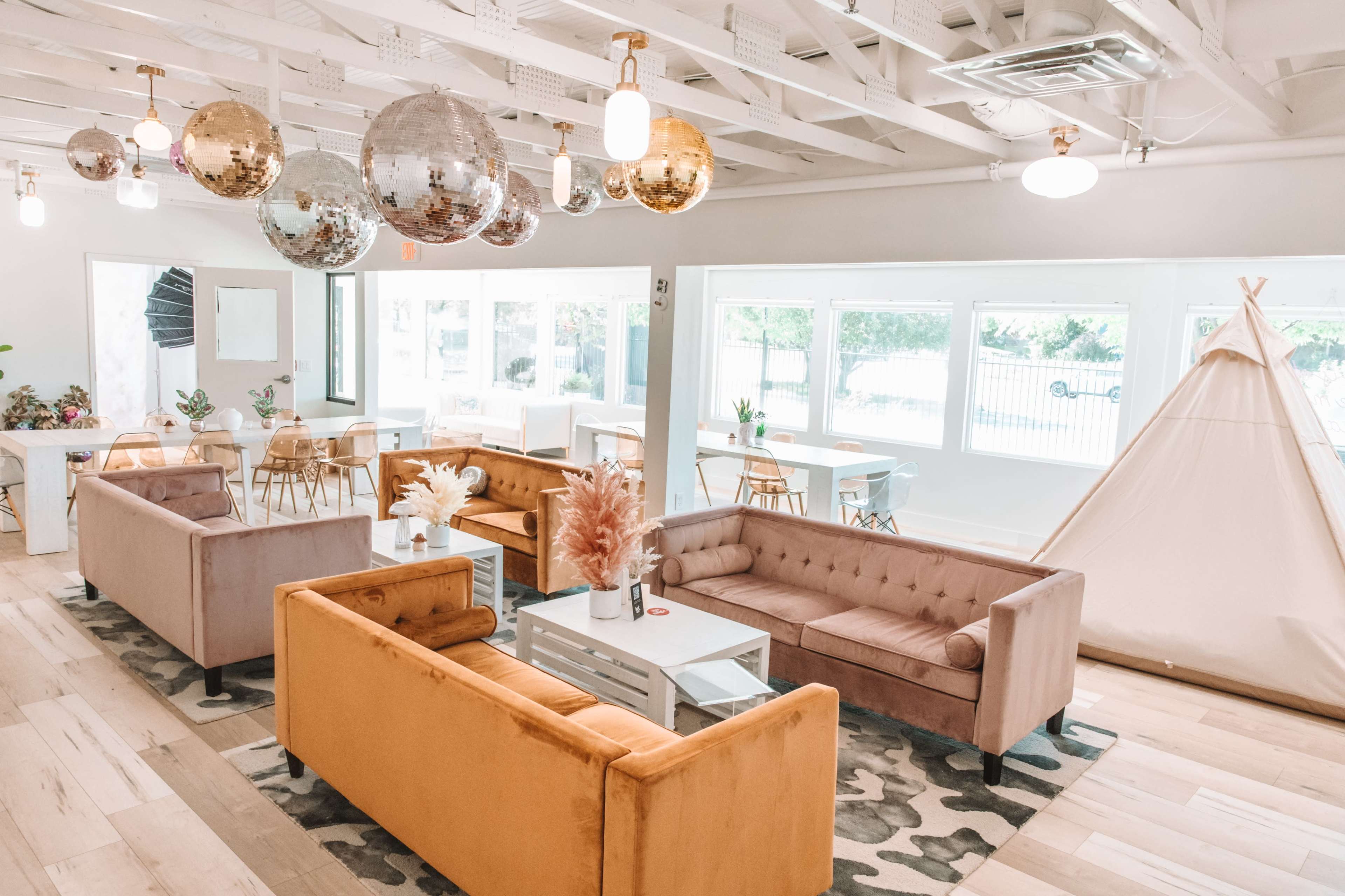A modern lounge area featuring plush sofas, hanging disco balls, a teepee, and large windows allowing natural light to fill the space.