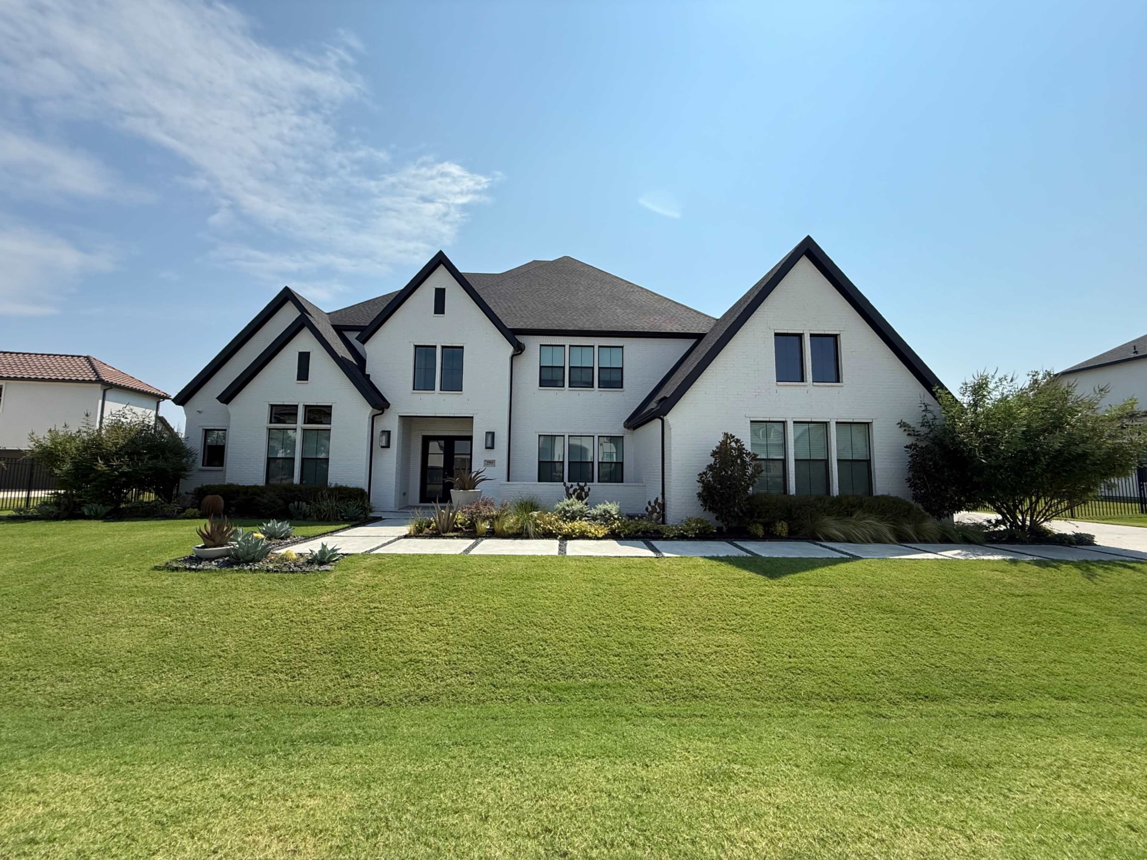 A large two-story white house with a black roof is set on a grassy lawn, featuring multiple windows and landscaped gardens in the front.